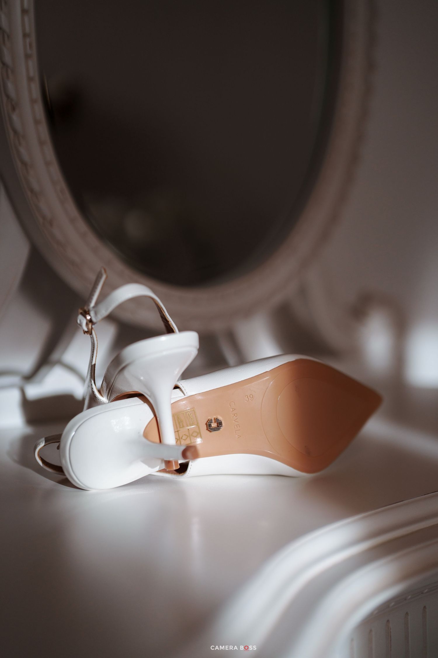White bridal high heel sandals with ankle strap resting beside a round mirror on a white vanity table.