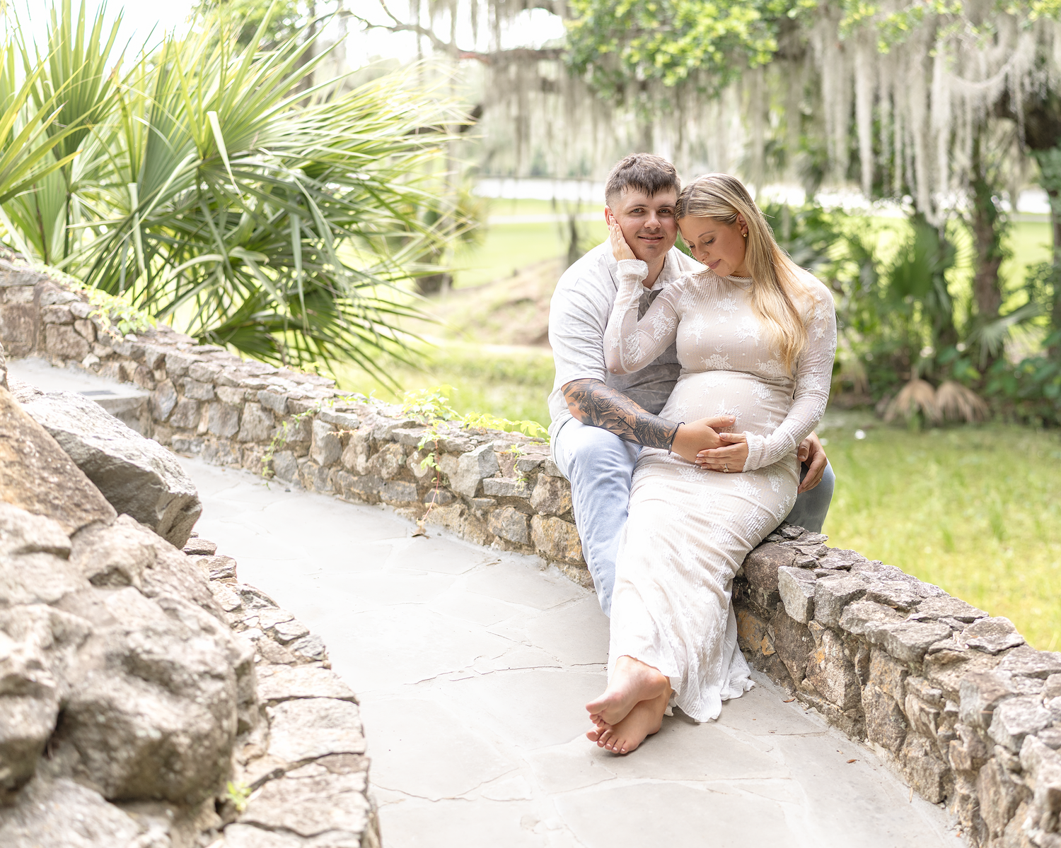 Couple sitting on a stone wall surrounded by lush greenery, woman in white lace maternity gown.