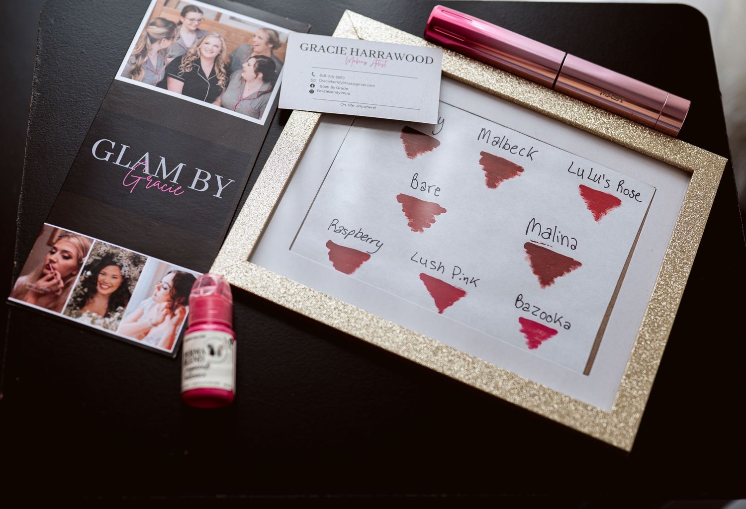 Glam By makeup artist branding display with lip color swatches, business cards, and cosmetic products on a glittery gold frame.