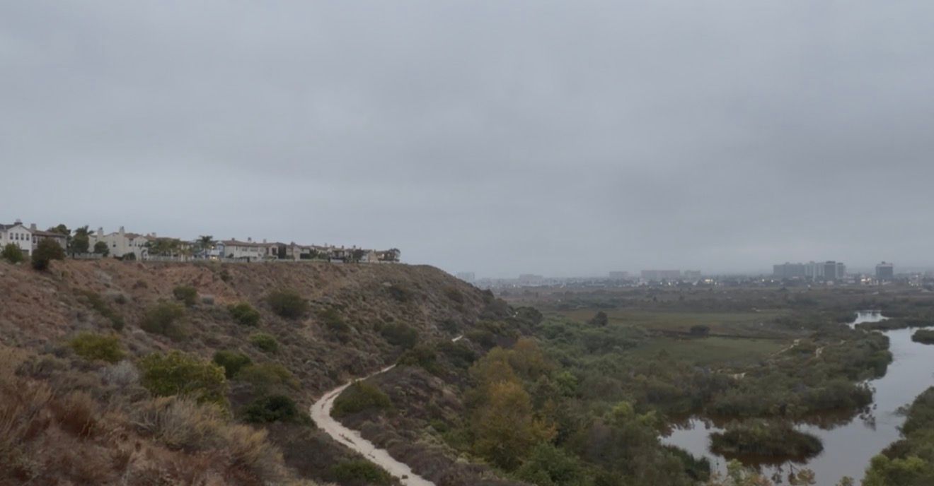 Panoramic view of a coastal wetland valley with winding trails, residential buildings, and a river under overcast skies.