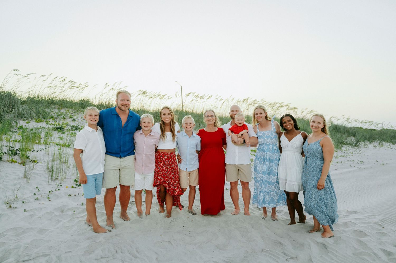 Large family group smiling together on a white sand beach with sea oats, wearing coordinated outfits.