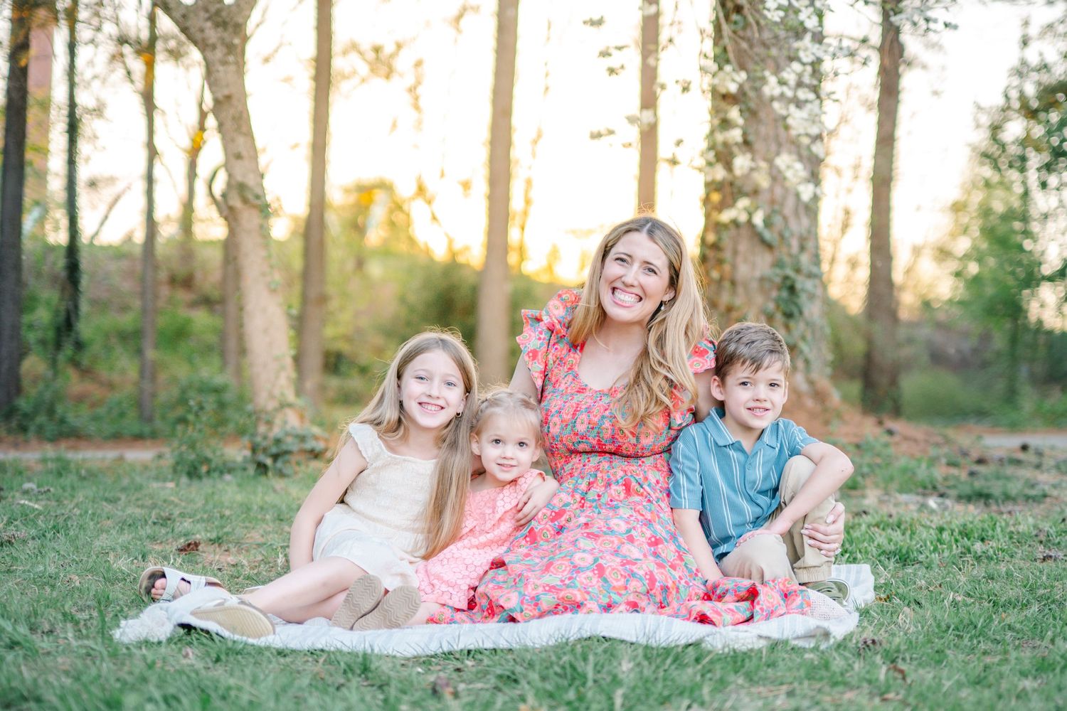 A smiling mother sits on a blanket in a sunny field with her three young children.