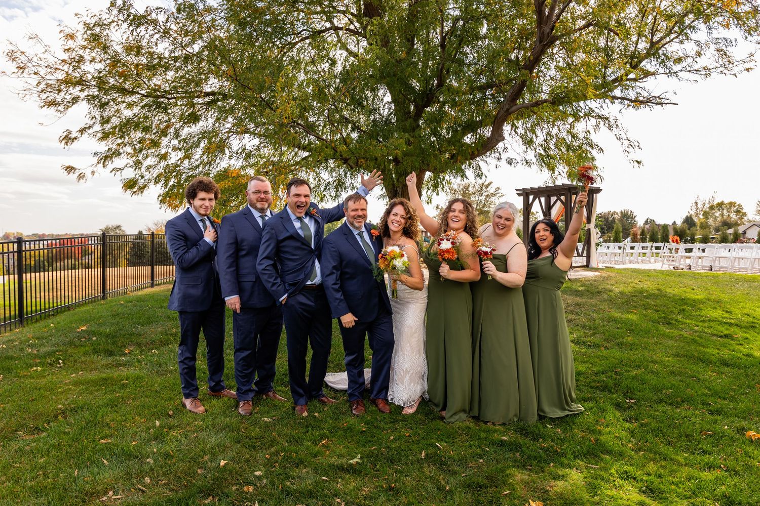 Wedding party posing outdoors under tree with bridesmaids in sage green dresses and groomsmen in navy at a Venue on the Ridge in Elkhorn, NE