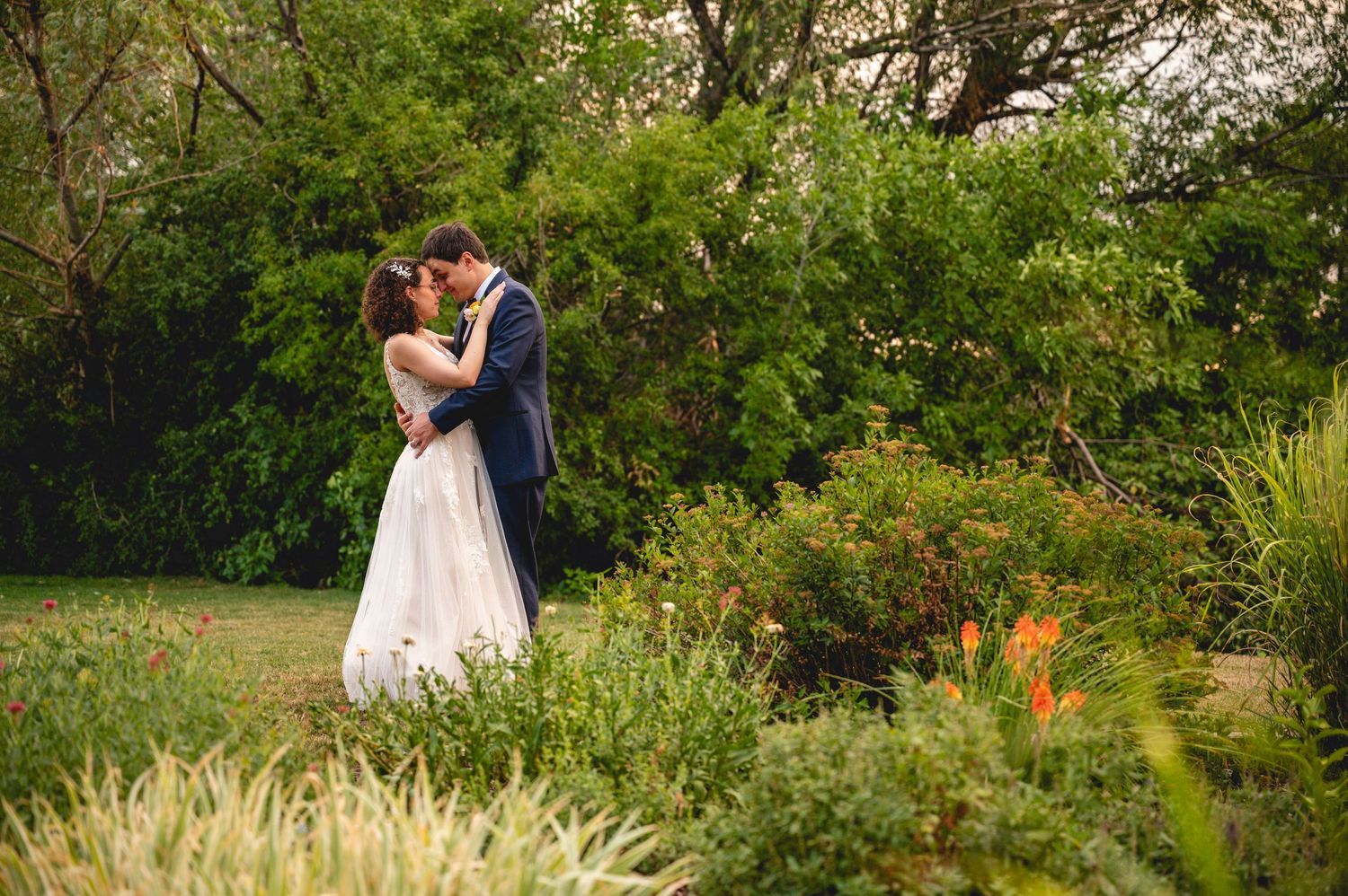 A bride and groom share a romantic kiss surrounded by lush green gardens on their wedding day.