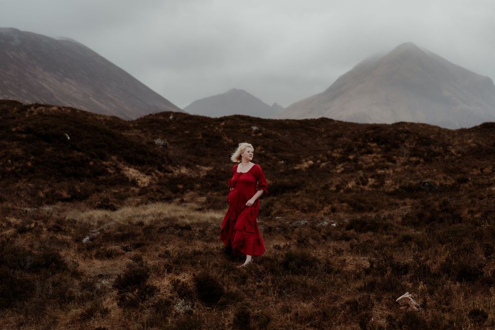 Woman in red dress walking through misty Scottish Highlands with dramatic mountain backdrop.