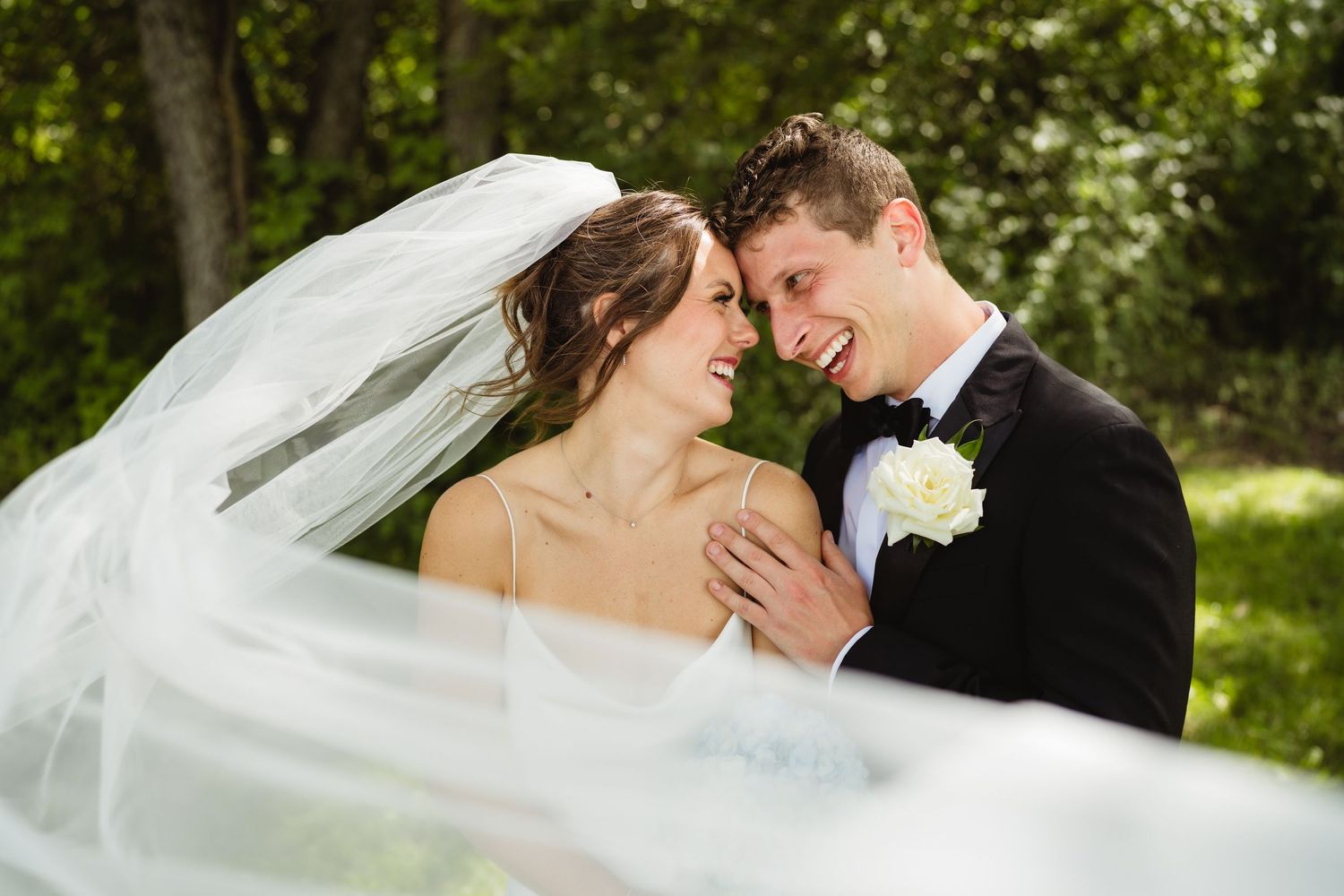 Bride and groom smiling together in a lush green meadow, groom in black tuxedo with boutonniere.