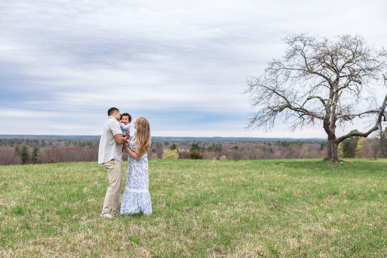 Husband and wife snuggle their baby boy in an open field with a bare tree in background on a cloudy day at Ward Hill Andover for family pics