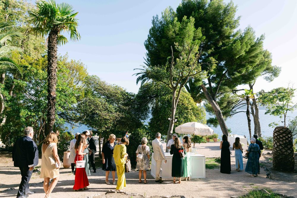 Elegant outdoor wedding reception with guests mingling among palm trees and lush greenery near a scenic waterfront venue.