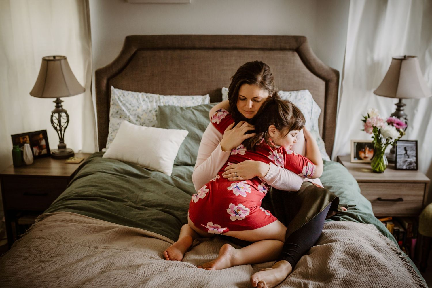 A mother sits on a bed embracing young child in a cozy bedroom with soft lighting.