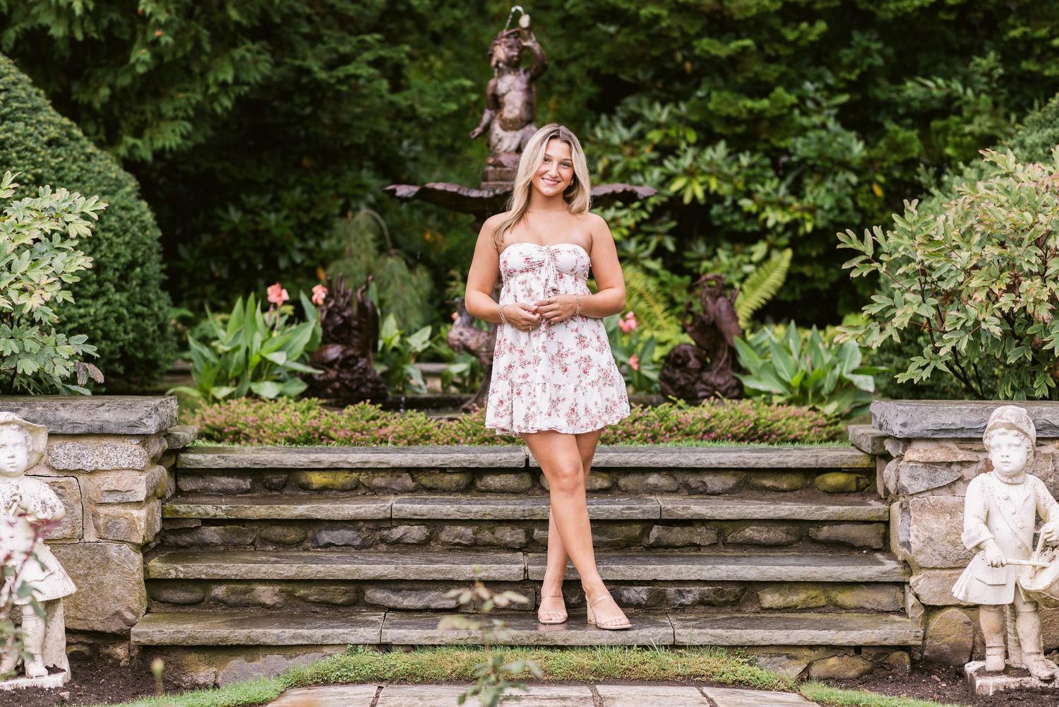 A blond senior girls poses in front of stone steps in a garden with greenery all around for senior photos at Fuller Gardens in Hampton, NH.