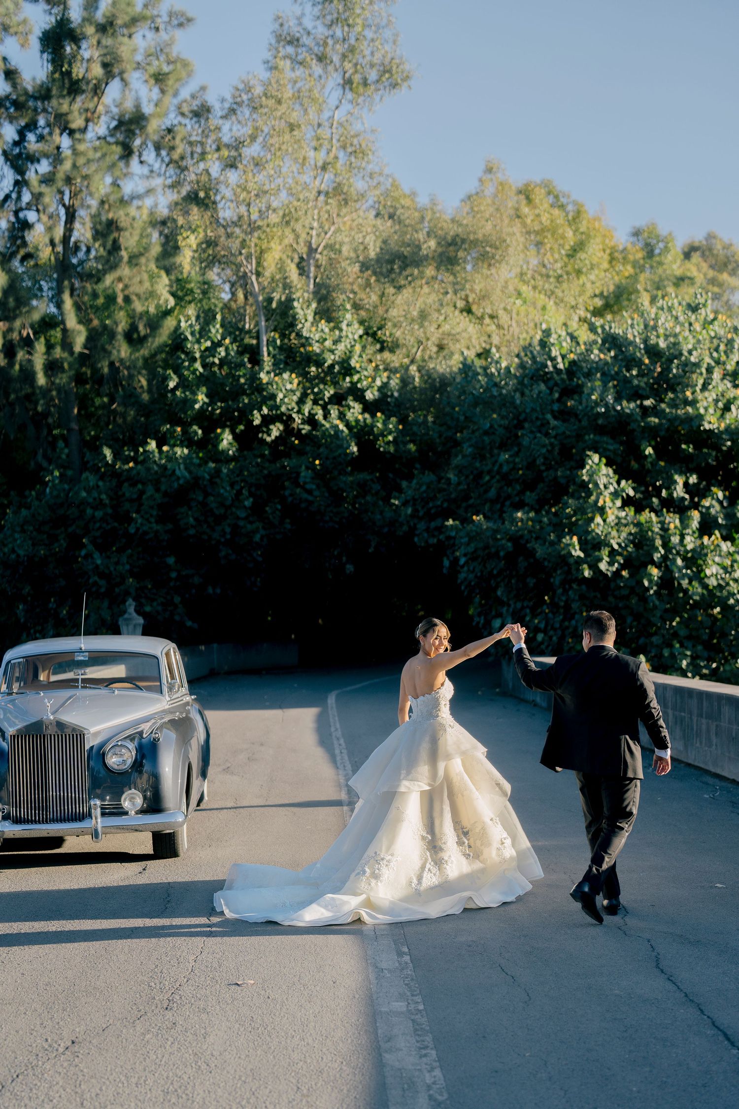 A couple in wedding attire walks and dances together on a paved path surrounded by lush greenery.