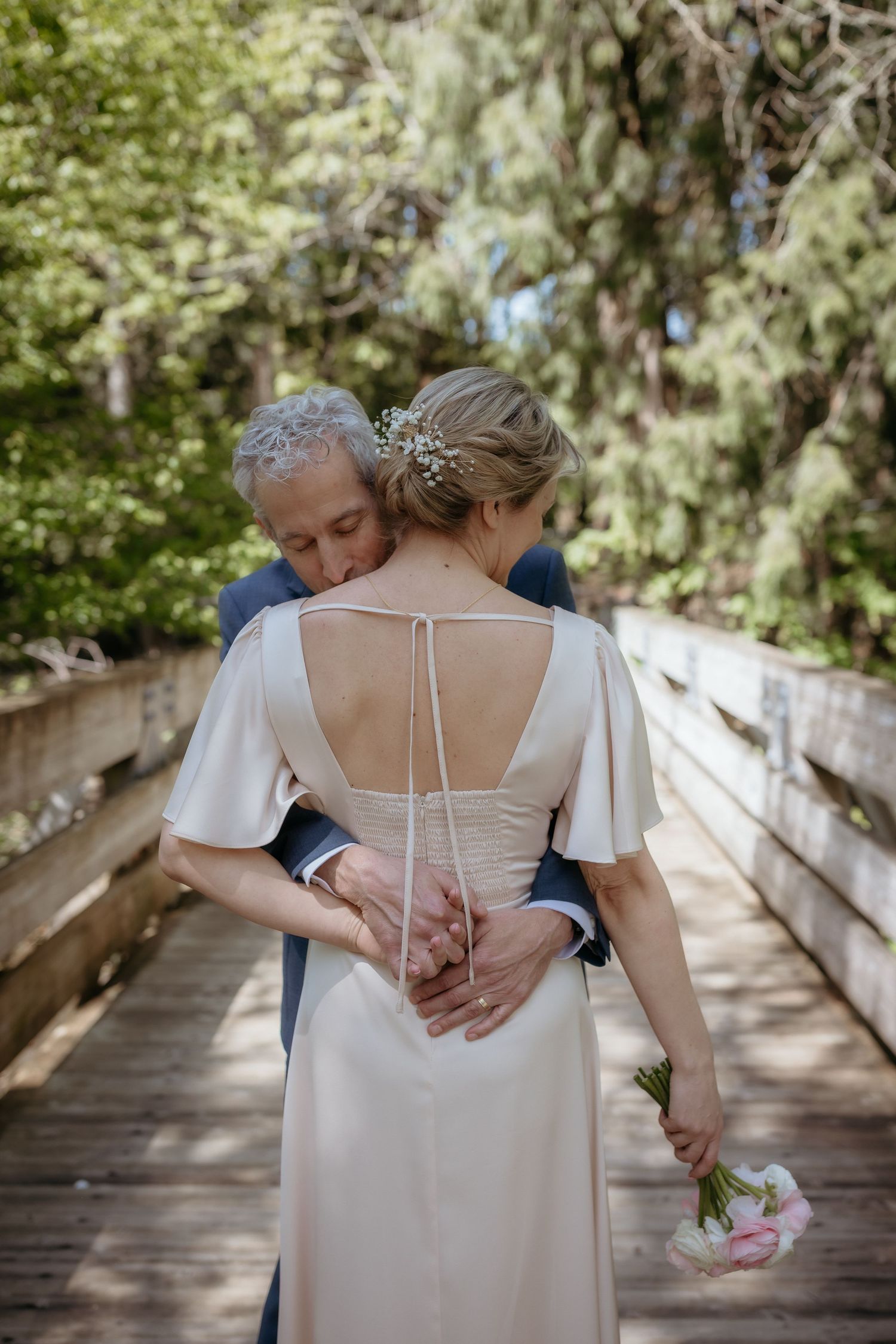 Bride and groom embracing on wooden bridge, bride in backless white dress holding pink bouquet, surrounded by lush green trees.