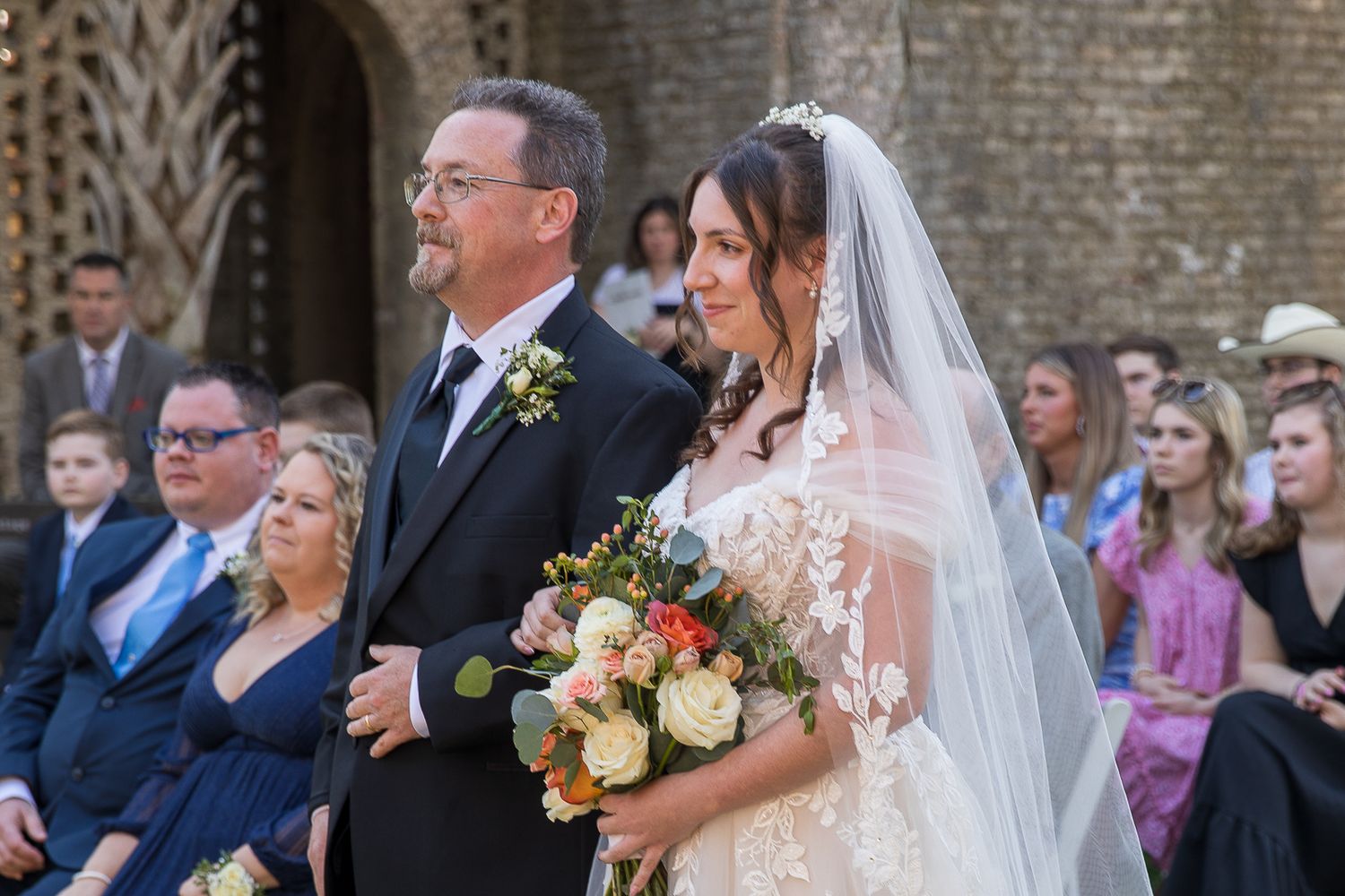 Bride in lace gown and veil holding floral bouquet walks down the aisle with escort at outdoor wedding ceremony.