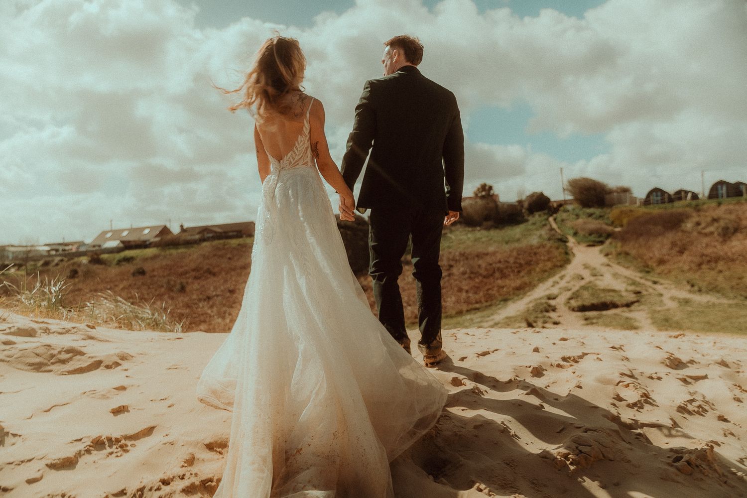 Bride and groom walking hand in hand on sandy beach dunes, wedding dress flowing in the wind on a cloudy day.