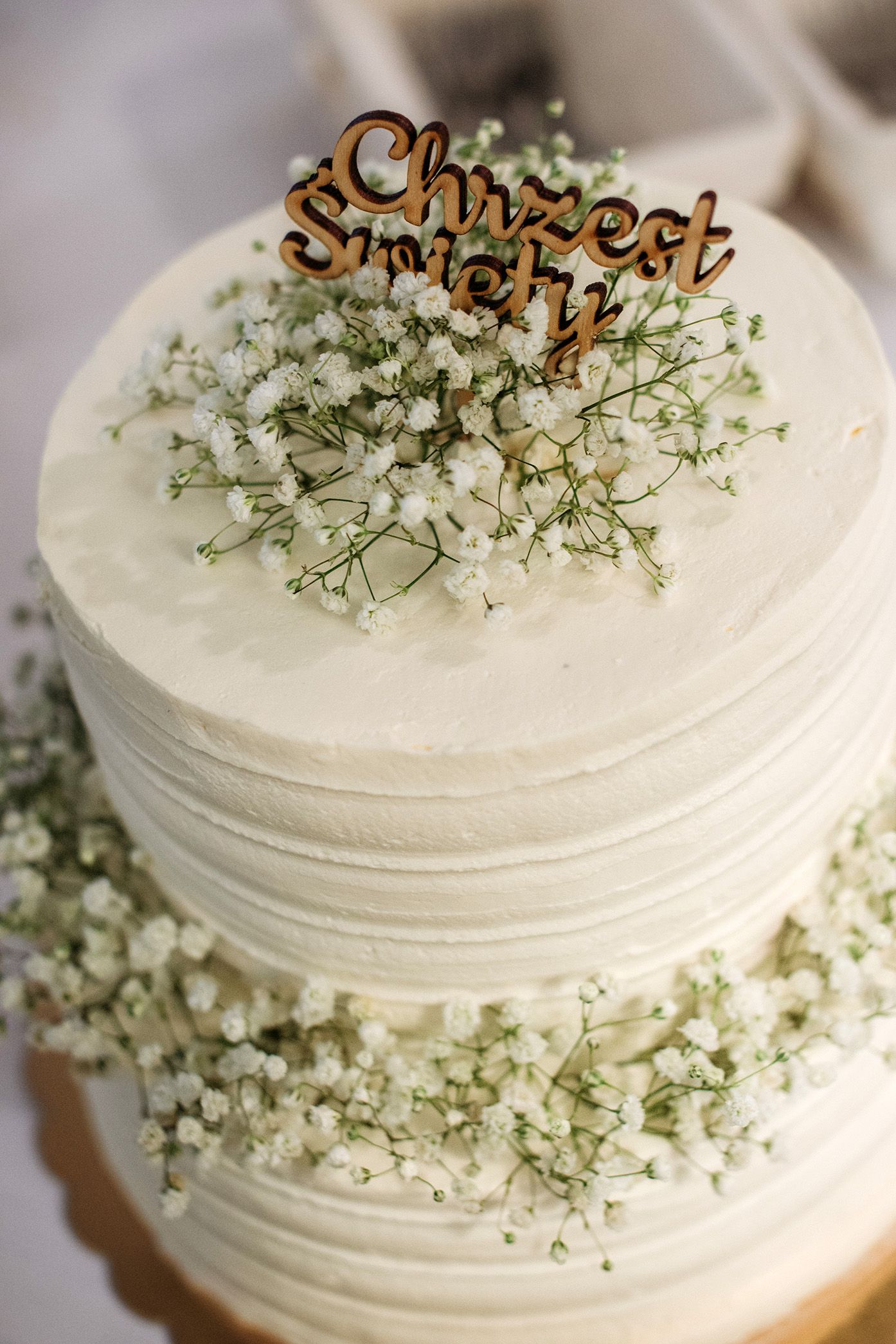 White wedding cake decorated with delicate baby's breath flowers and a rustic wooden topper against a light background.