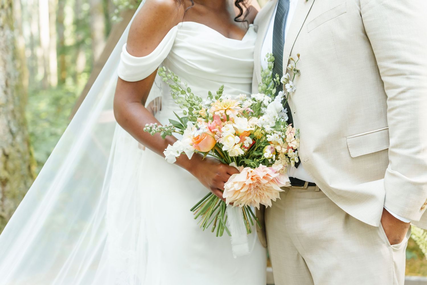 Bride in off-shoulder white gown holding peach floral bouquet beside groom in beige suit outdoors.