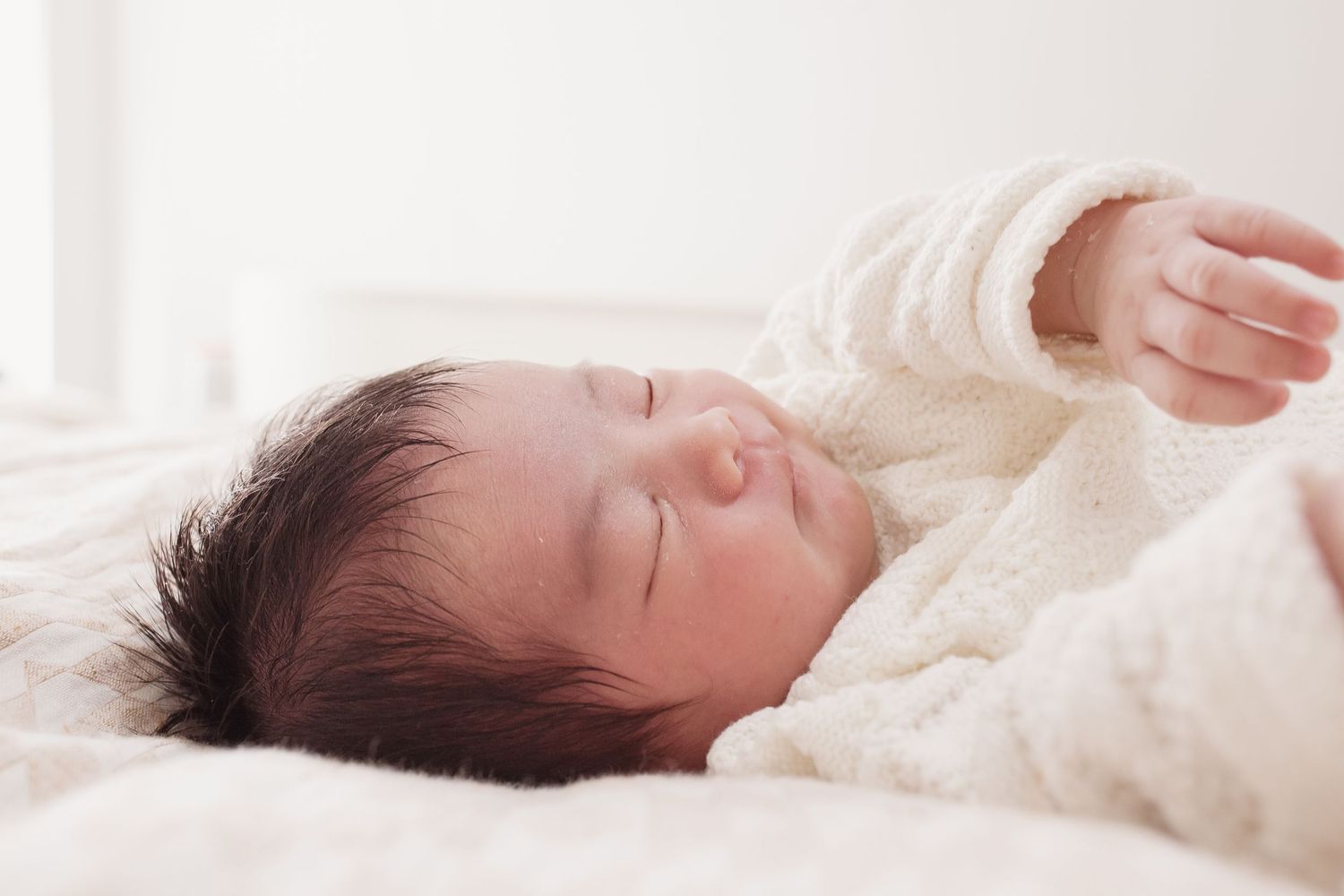 Sleeping baby in white sweater stretching on soft bedding.