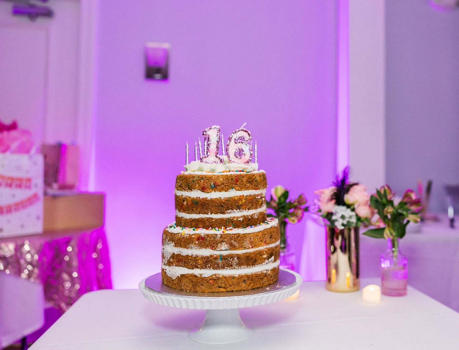 A three-tier naked birthday cake with '16' candles on a white stand, set against purple ambient lighting.