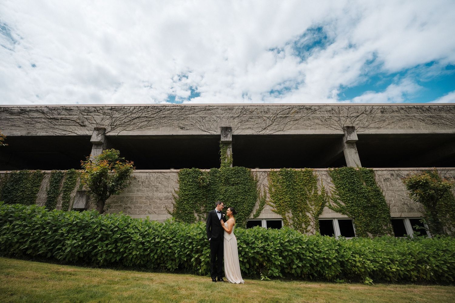 Couple embraces on lush green lawn before ivy-covered stone wall under dramatic cloudy sky.