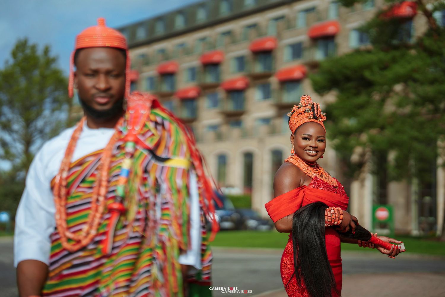 Couple in vibrant African traditional attire, man in kente robe and woman in red dress with orange headpiece, outdoors.