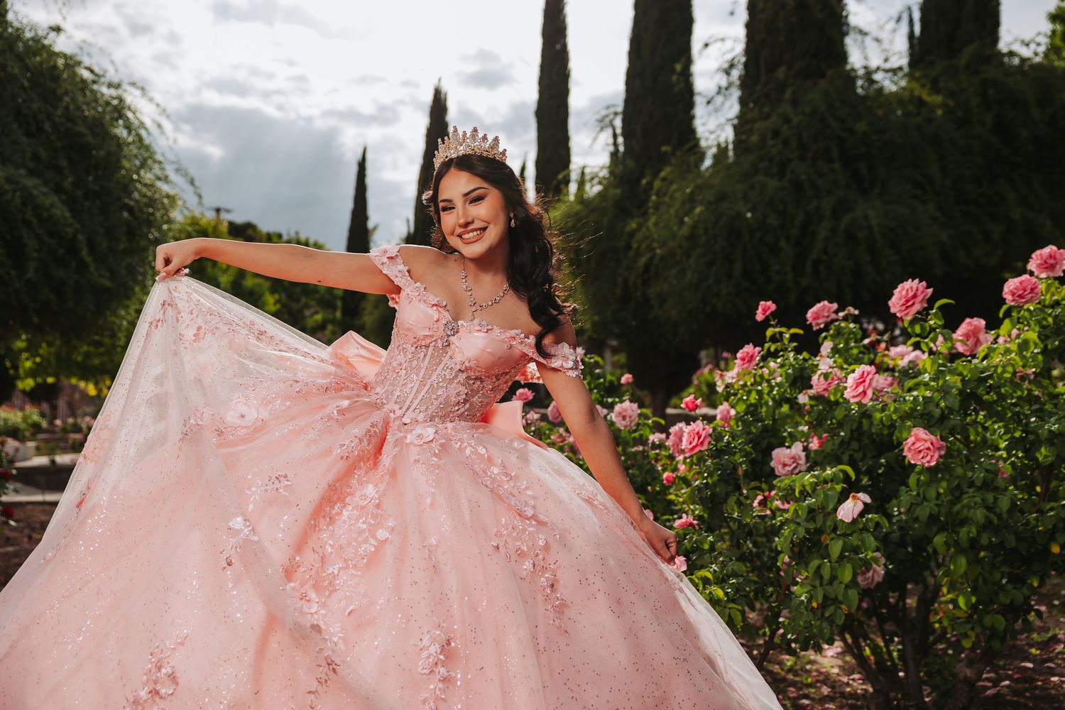 Young Latina girl in pink quinceañera ball gown with floral details and tiara poses in a rose garden.