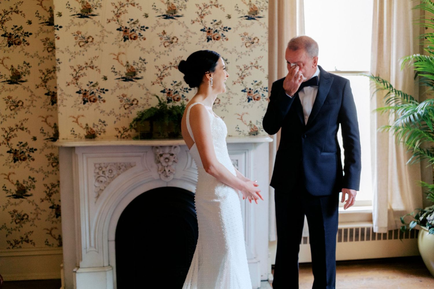Emotional father wipes tears seeing bride daughter in white wedding gown during first look moment by fireplace.