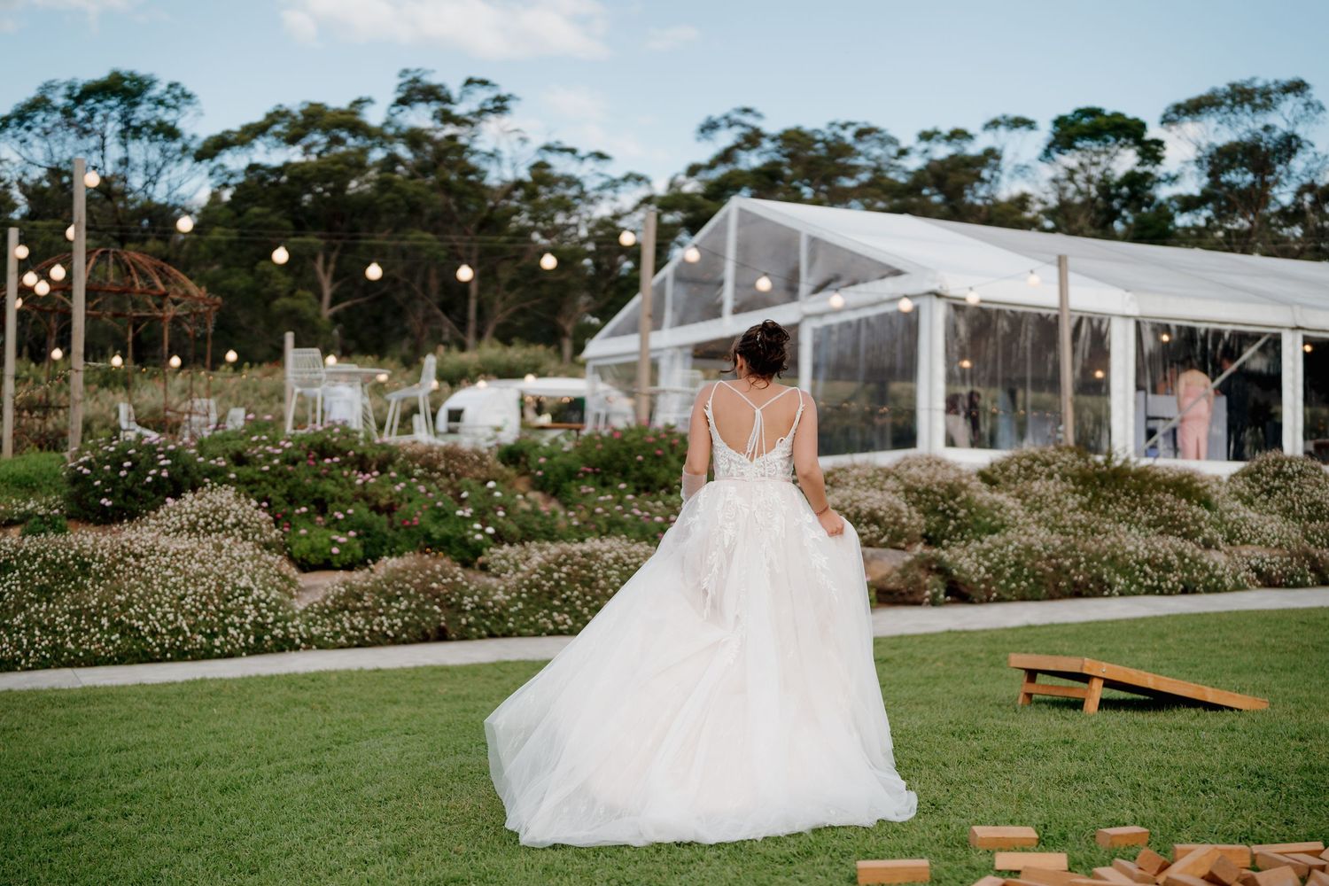 Bride in a stunning backless white gown stands on lush green lawn before a marquee wedding venue surrounded by gardens.