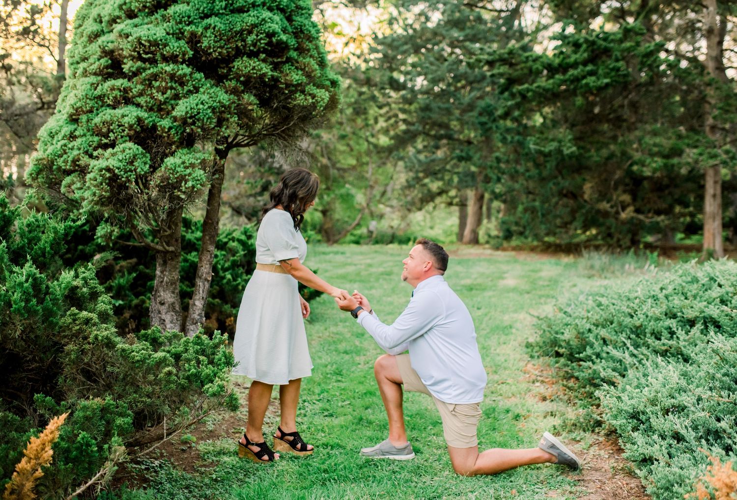 Man kneeling on one knee proposing to woman in white dress outdoors in lush green garden setting.