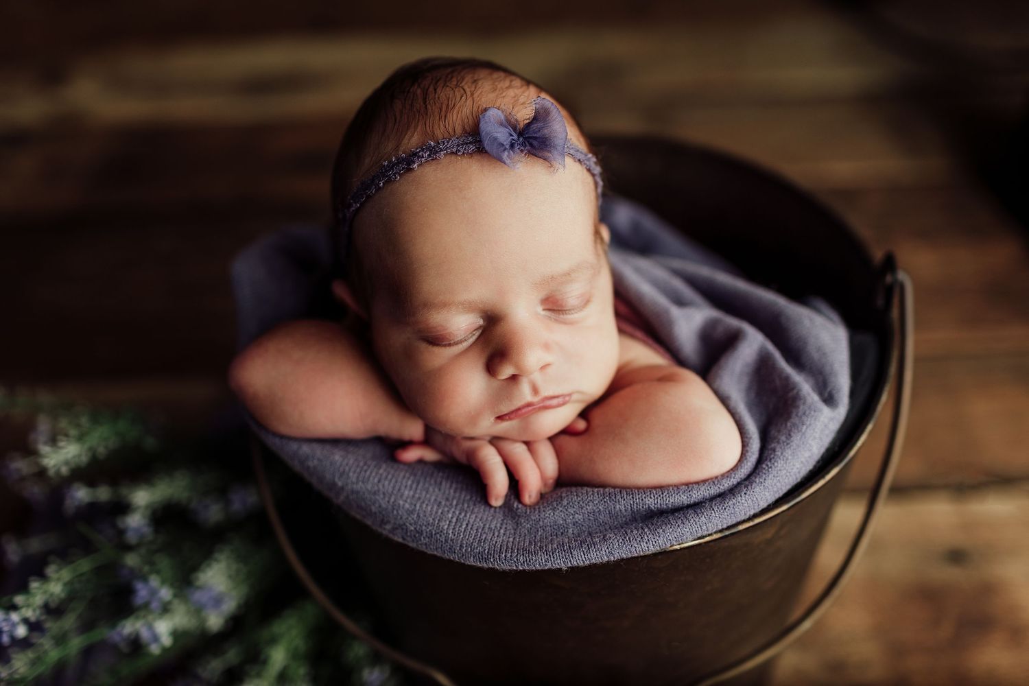 Sleeping newborn baby girl in a metal bucket with purple blanket and lavender bow headband on a wooden background.