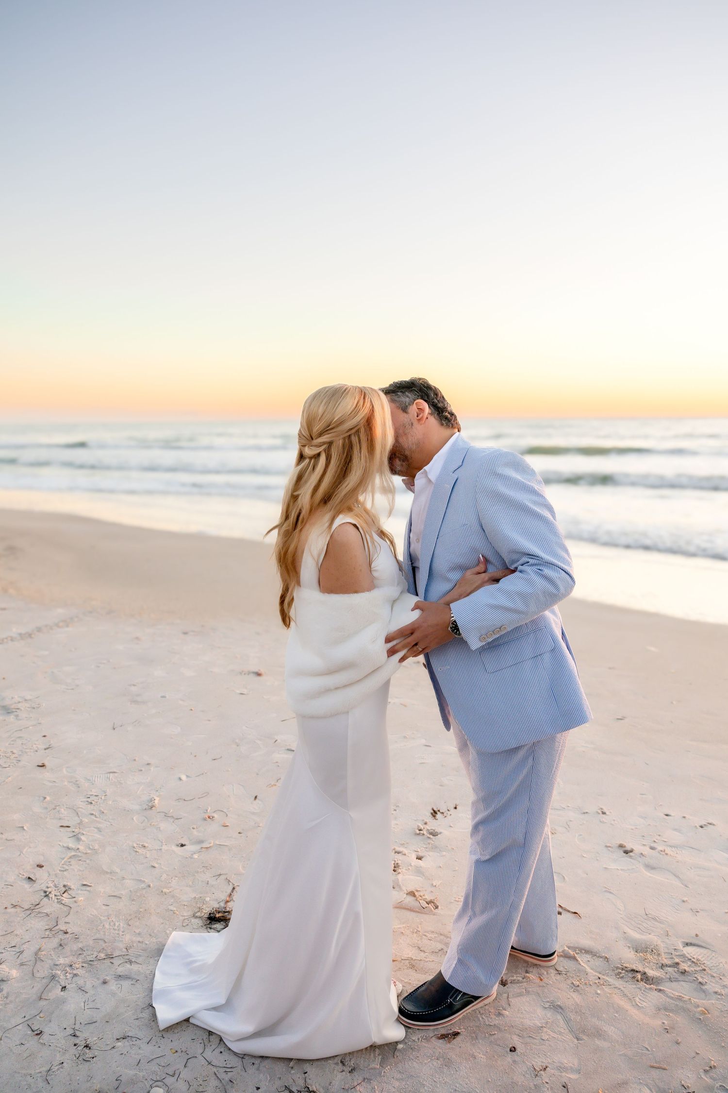 A bride and groom kiss on a sandy beach with a pastel sunset sky behind them.
