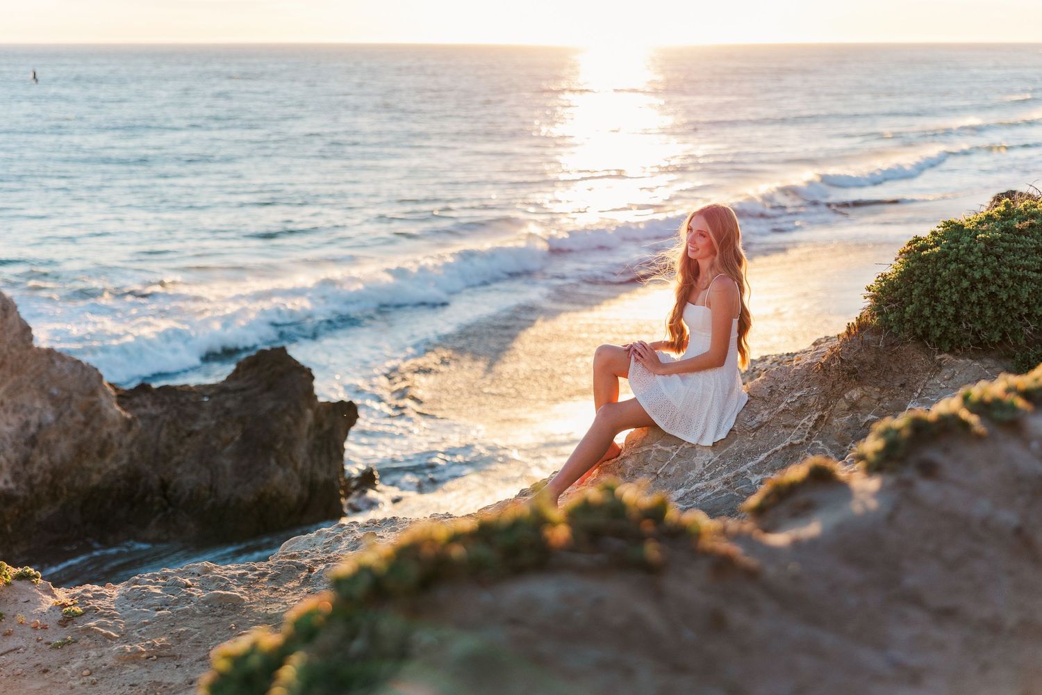 Woman in white dress poses gracefully on coastal cliffs during golden hour sunset by the ocean.