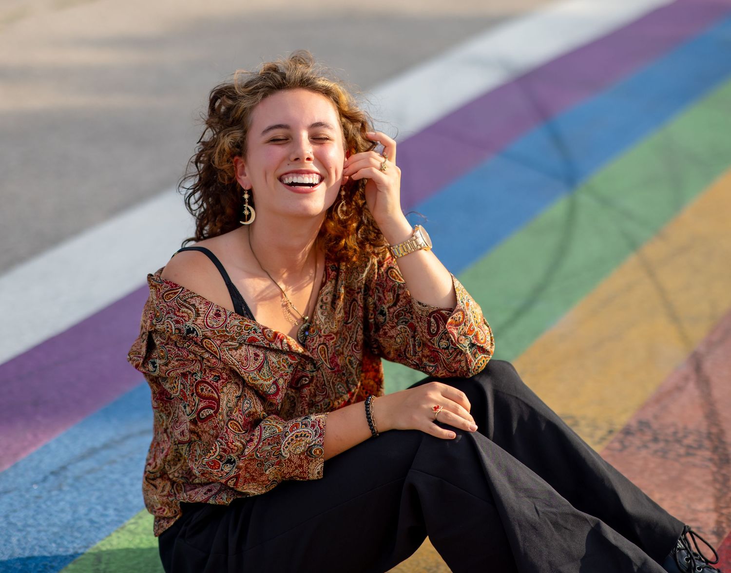 A person with curly hair wearing a floral dress poses joyfully on colorful rainbow painted steps outdoors.