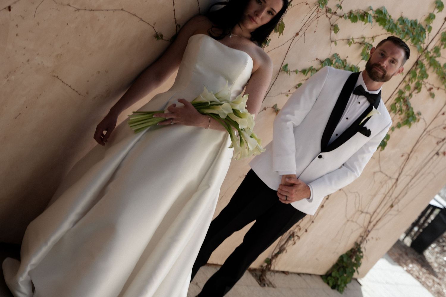 Bride and groom pose together in a lush garden beside a rustic venue, bride in white gown, groom in black tuxedo.