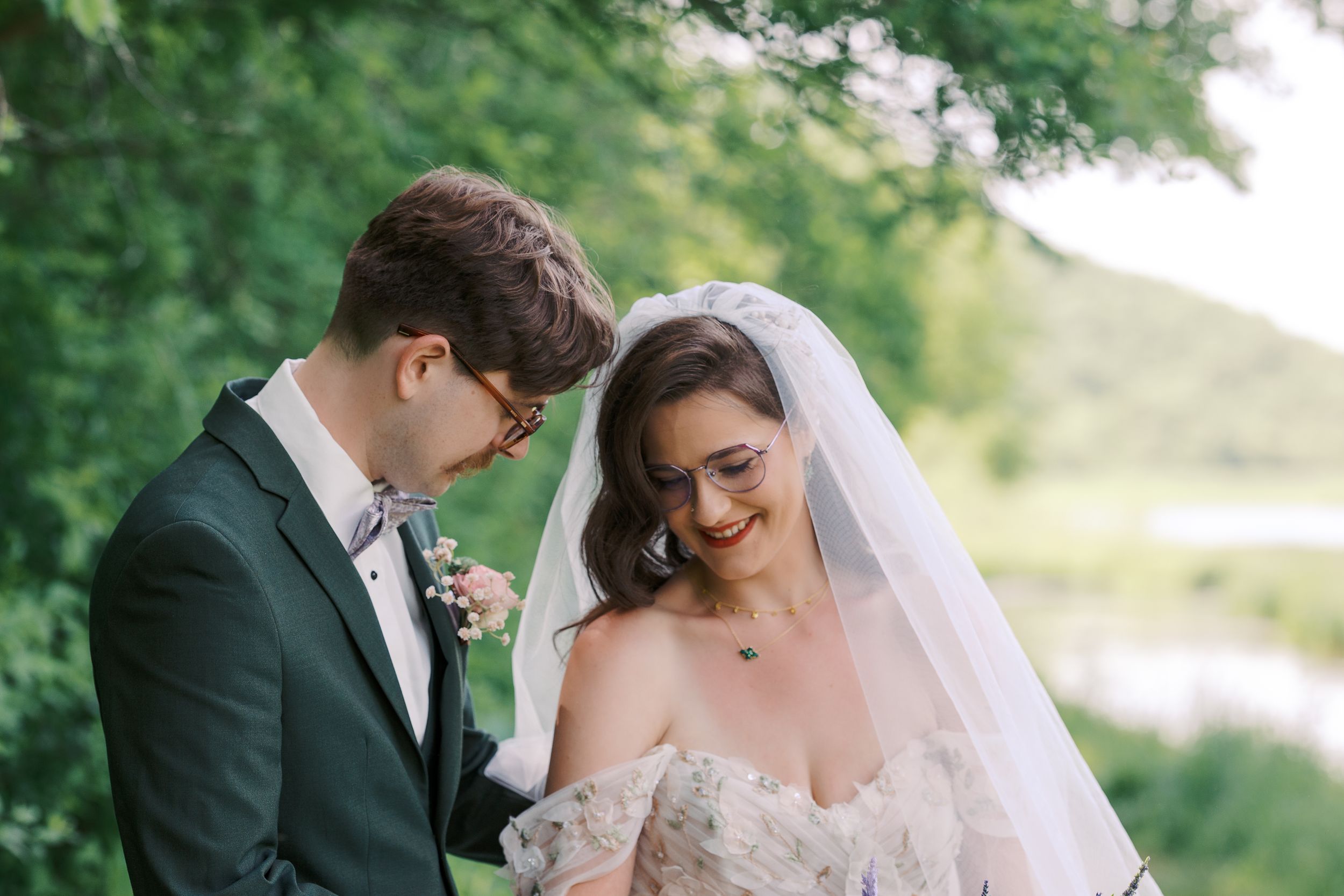 Bride and groom share a tender moment outdoors, surrounded by lush green trees, bride wearing a lace gown and veil.