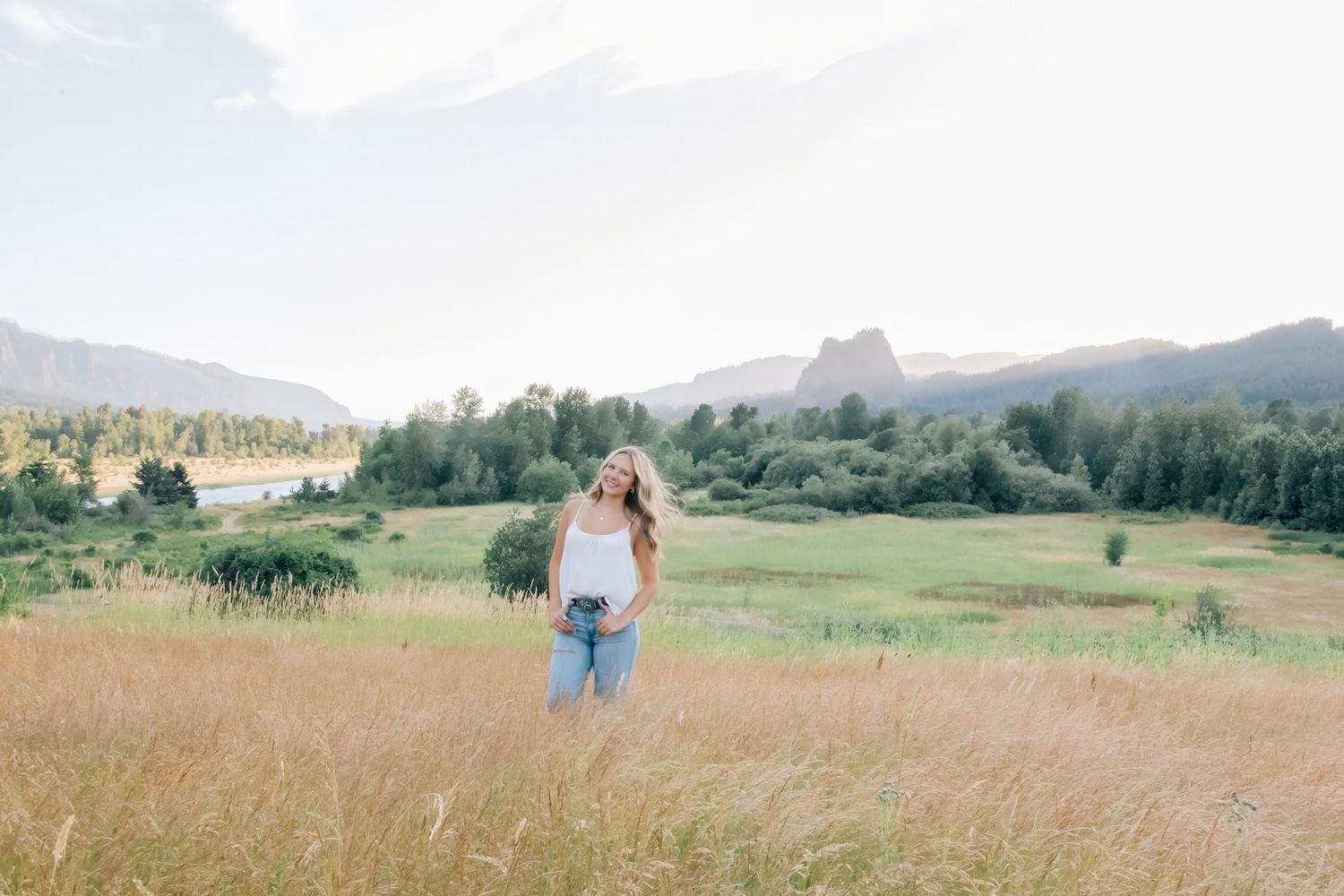 Woman standing in golden meadow with mountains, river, and green trees in scenic landscape background.