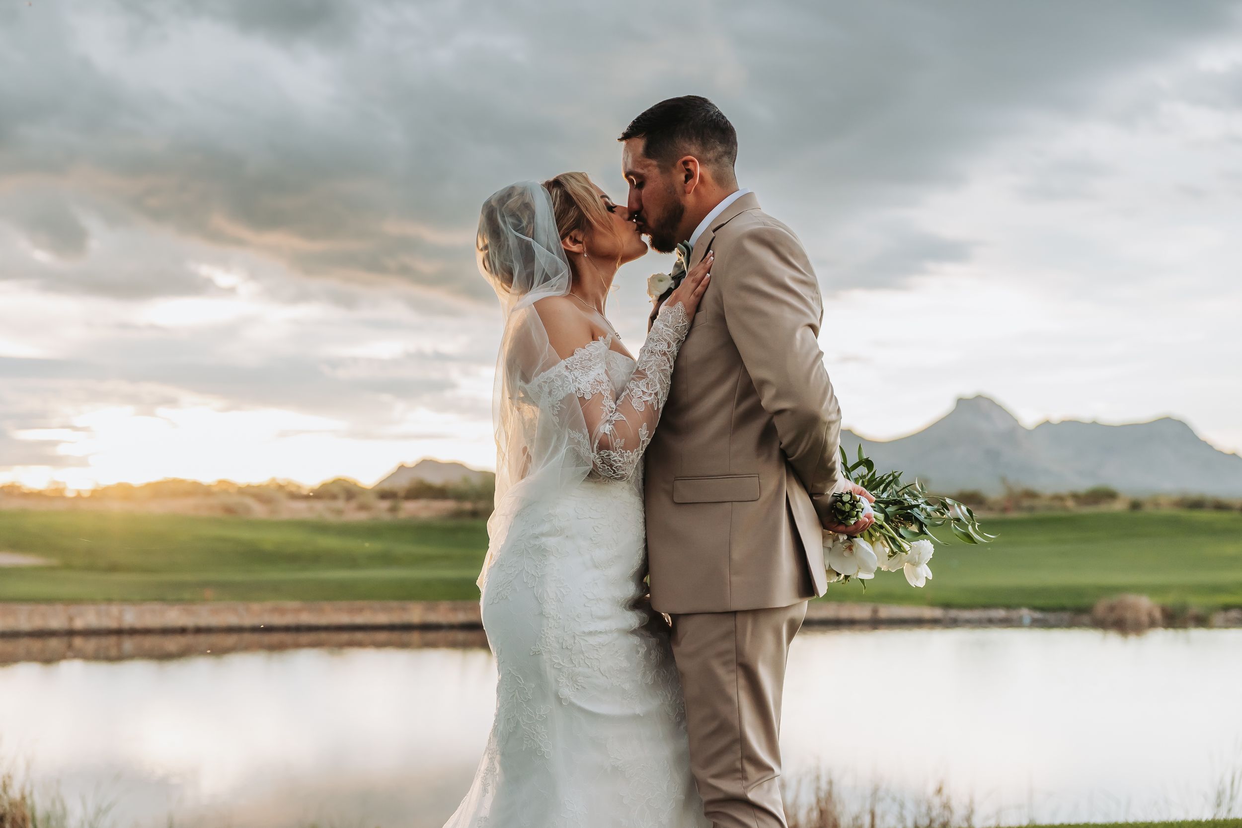 Bride and groom sharing a romantic kiss at sunset near a pond, with mountains in the background on their wedding day.