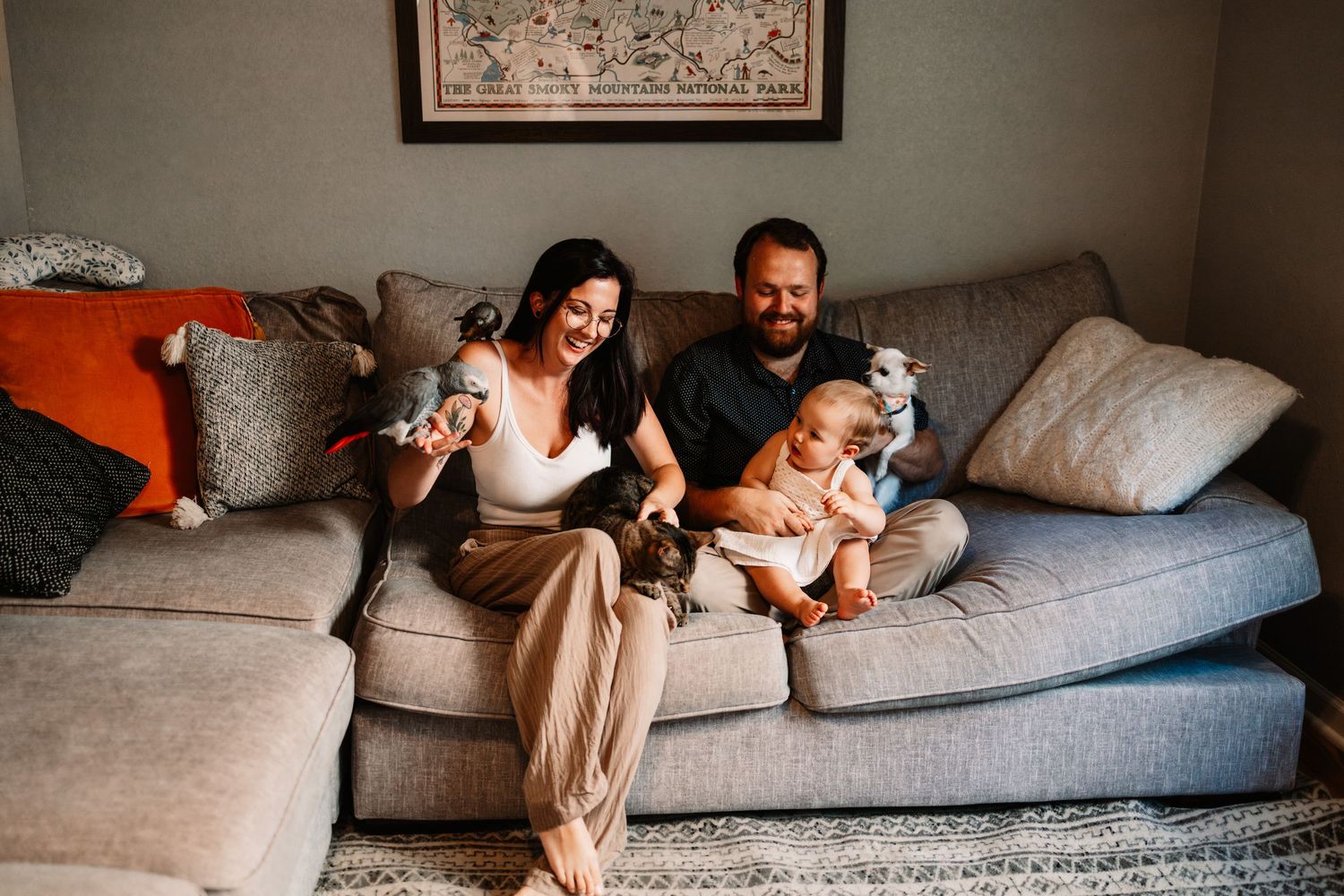 Happy family sitting on gray couch, parents smiling with baby and toddler in cozy living room in Knoxville, TN.