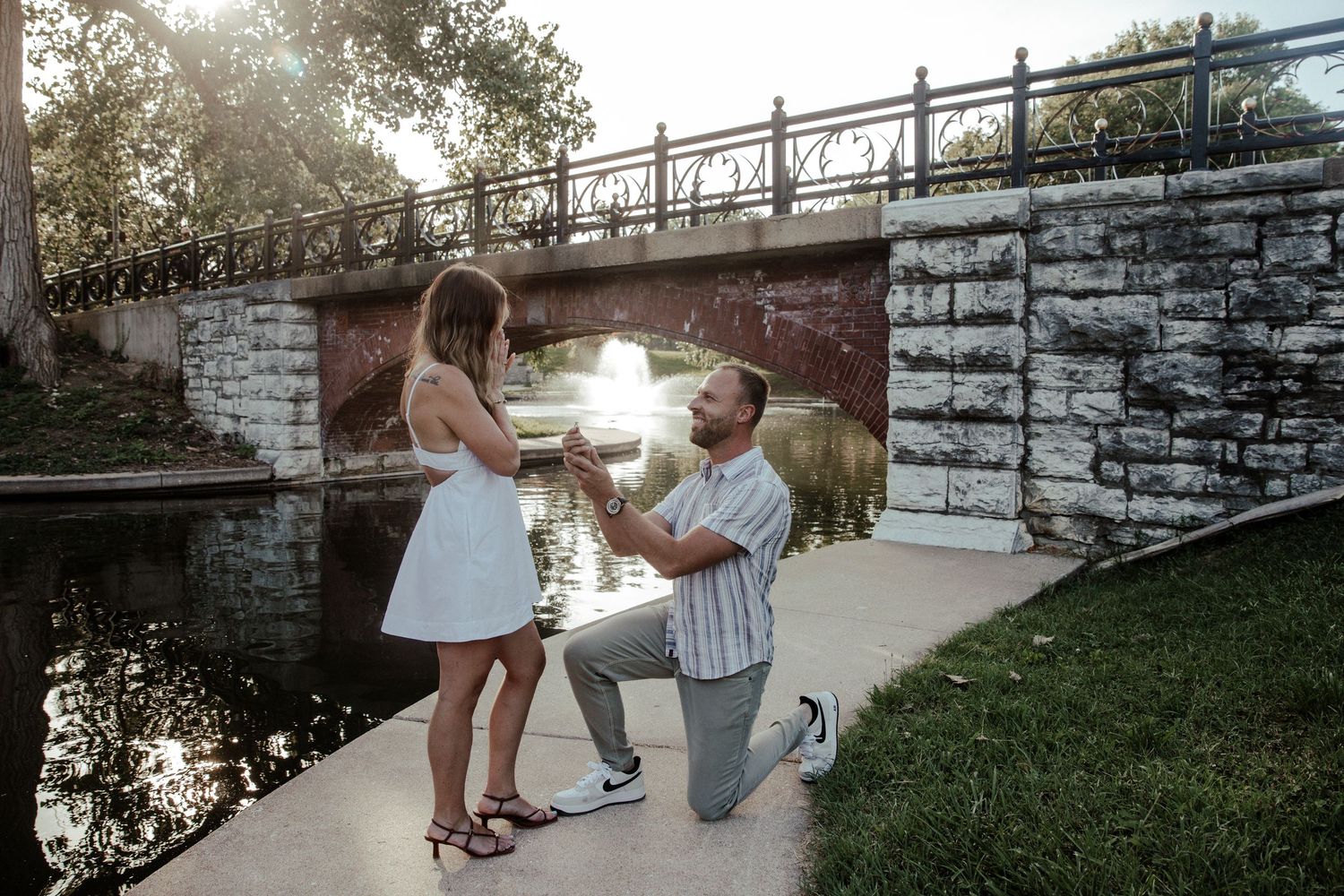 Man proposes to woman on one knee by a scenic bridge and canal at sunset, holding an engagement ring.
