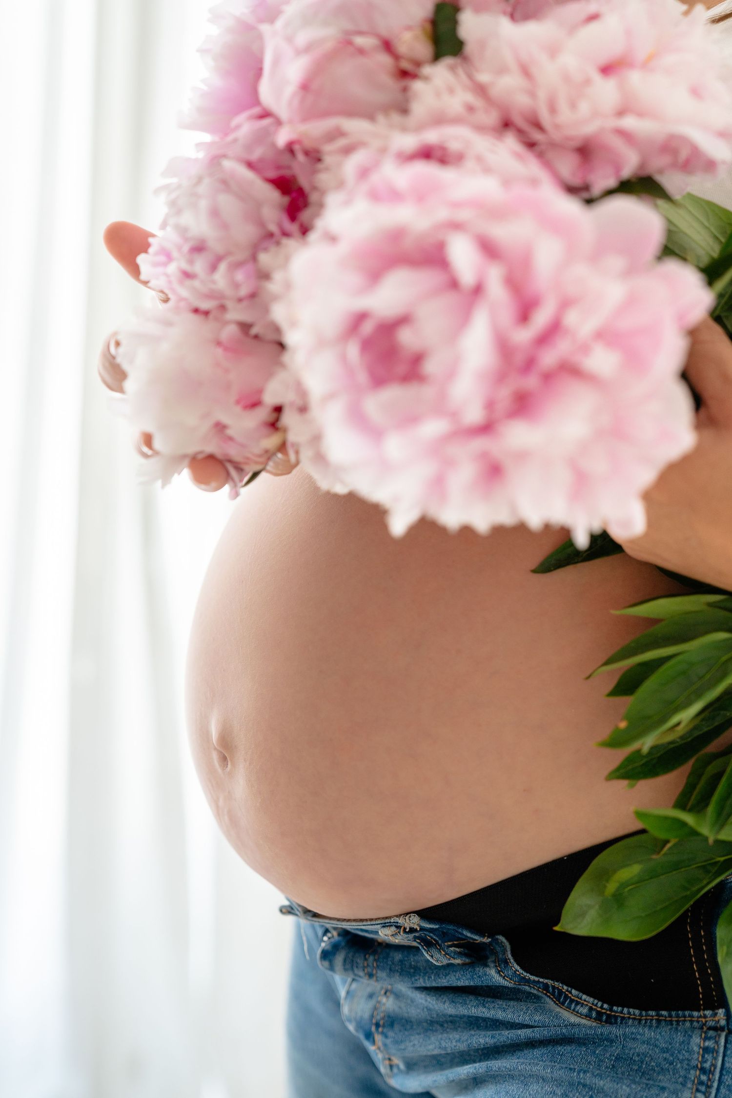 Pregnant woman holding pink peonies bouquet over bare baby bump, wearing jeans, standing near white curtains.