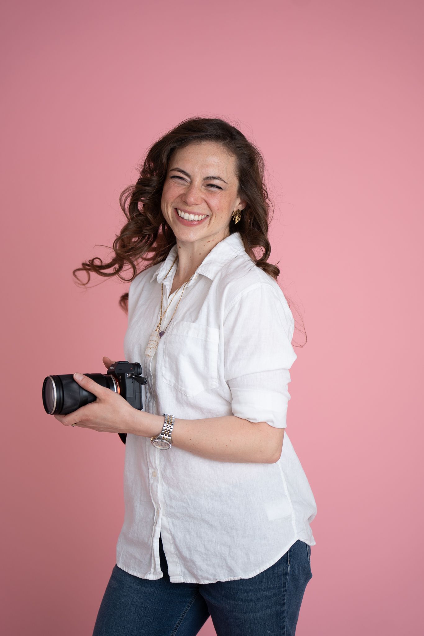 Woman in white shirt holding a DSLR camera on her shoulder, posing confidently against a tan background.