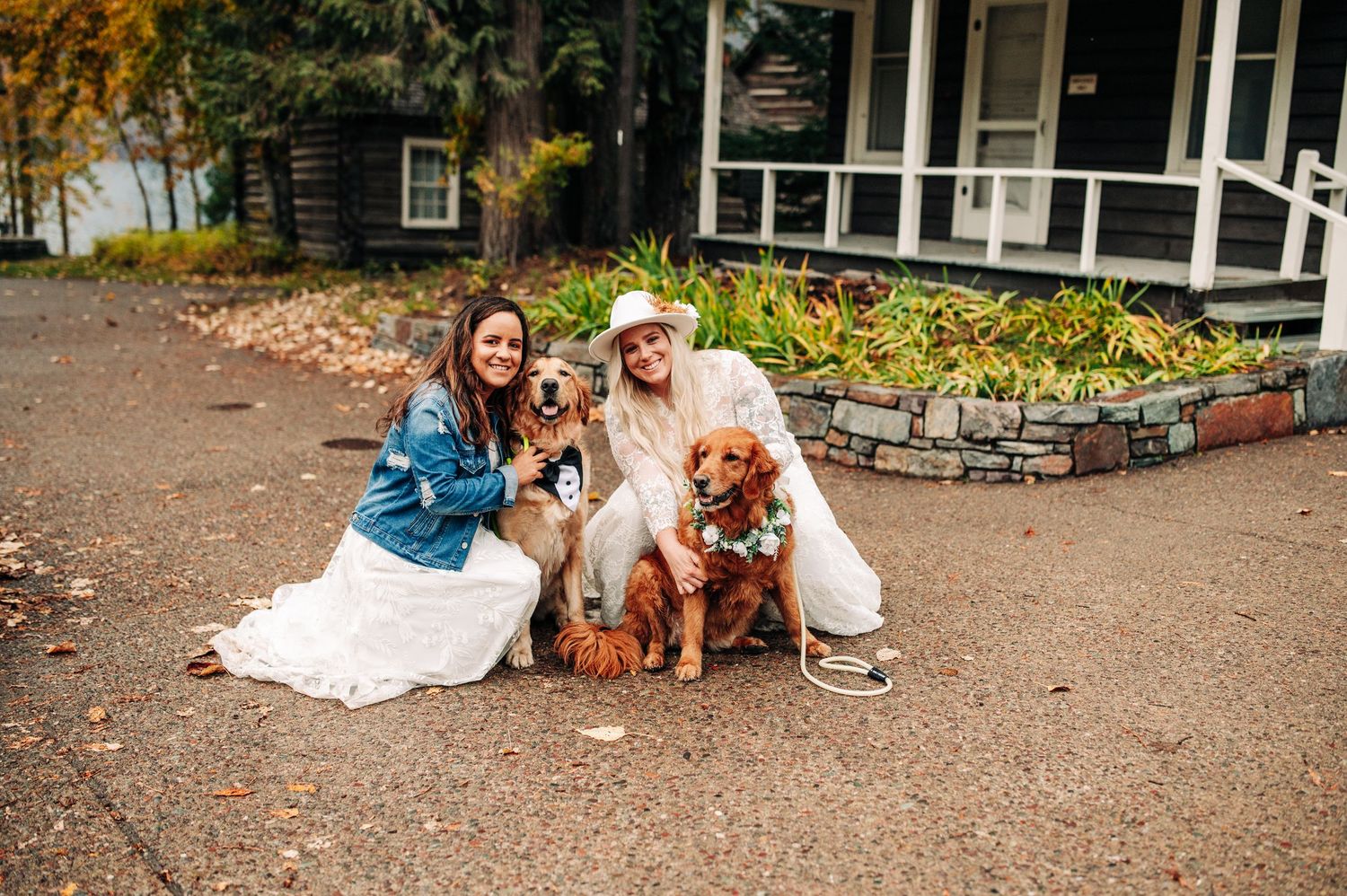 Two brides pose with golden retrievers outside a rustic venue during an autumn wedding celebration.