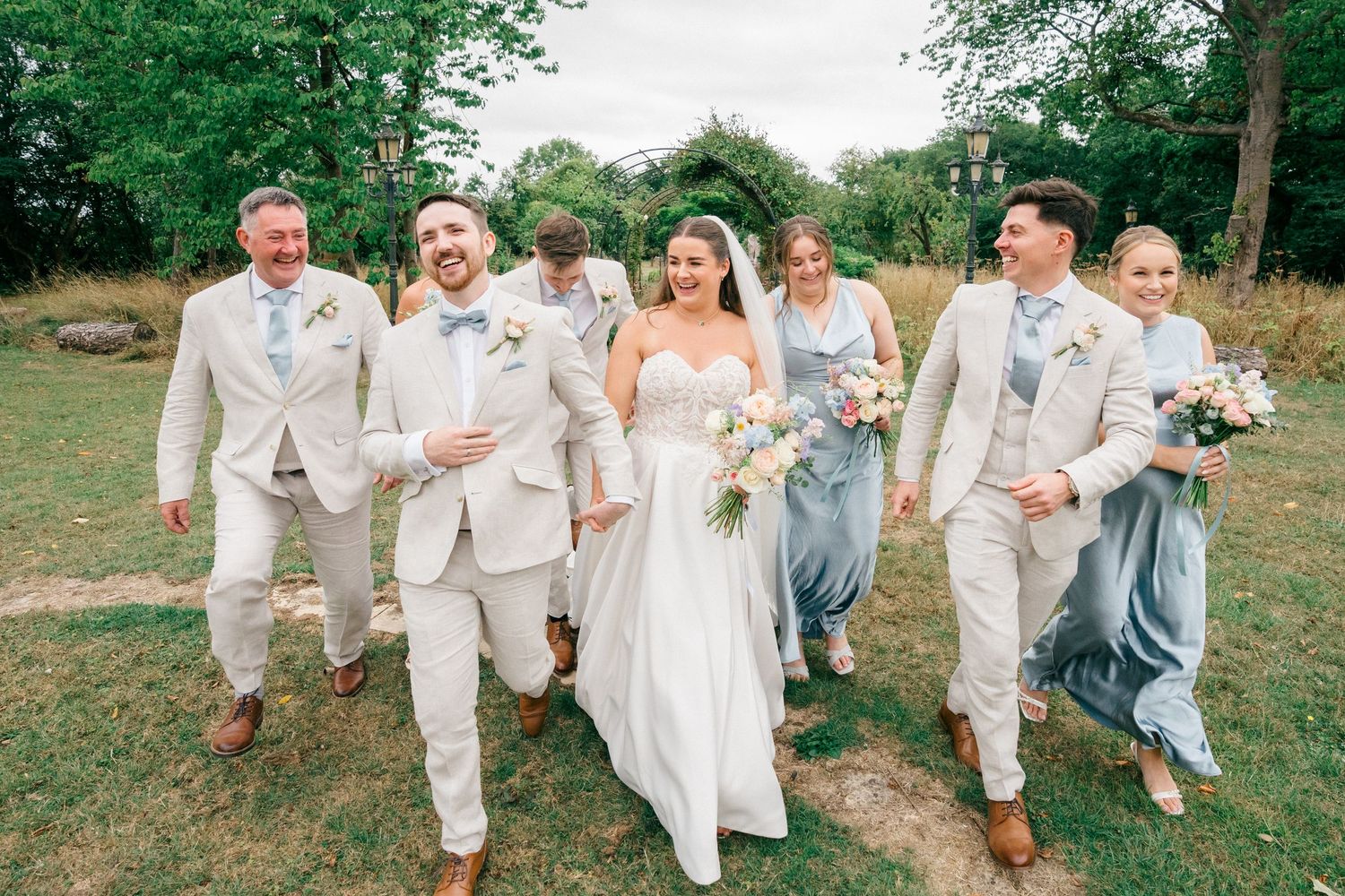 Wedding party walks outdoors in beige suits and blue bridesmaid dresses with bride in white gown holding bouquet.