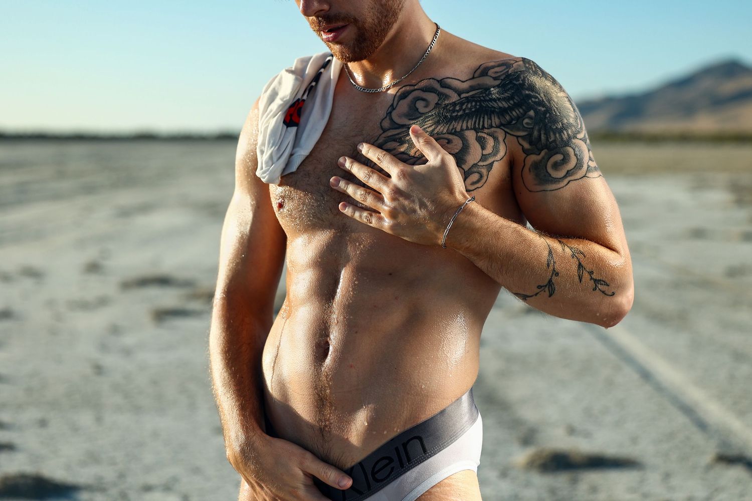 Muscular tattooed man in Calvin Klein underwear posing shirtless outdoors in a dry desert landscape.