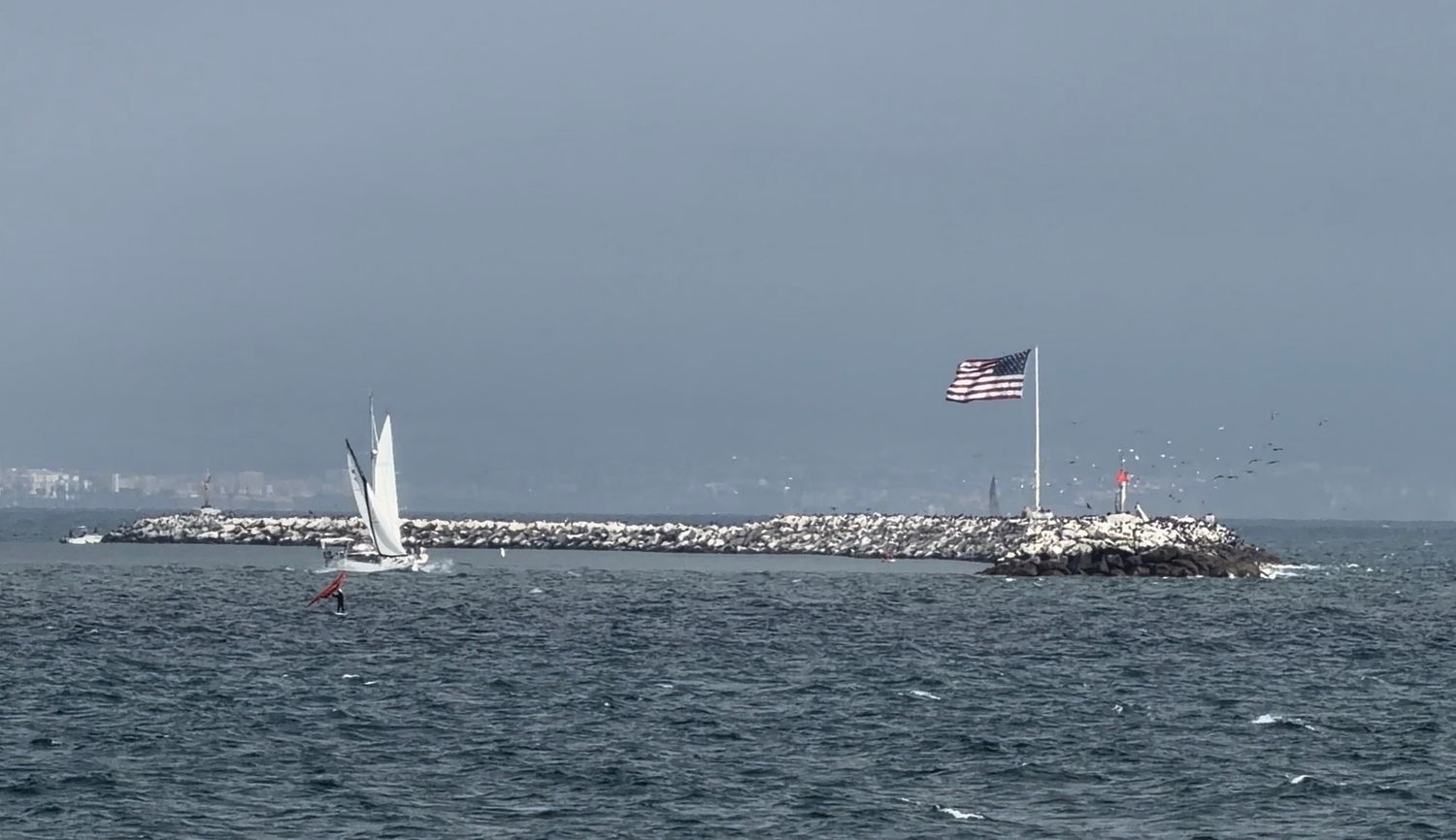 Sailboat navigating choppy ocean waters near a rocky breakwater with an American flag flying on a pole.