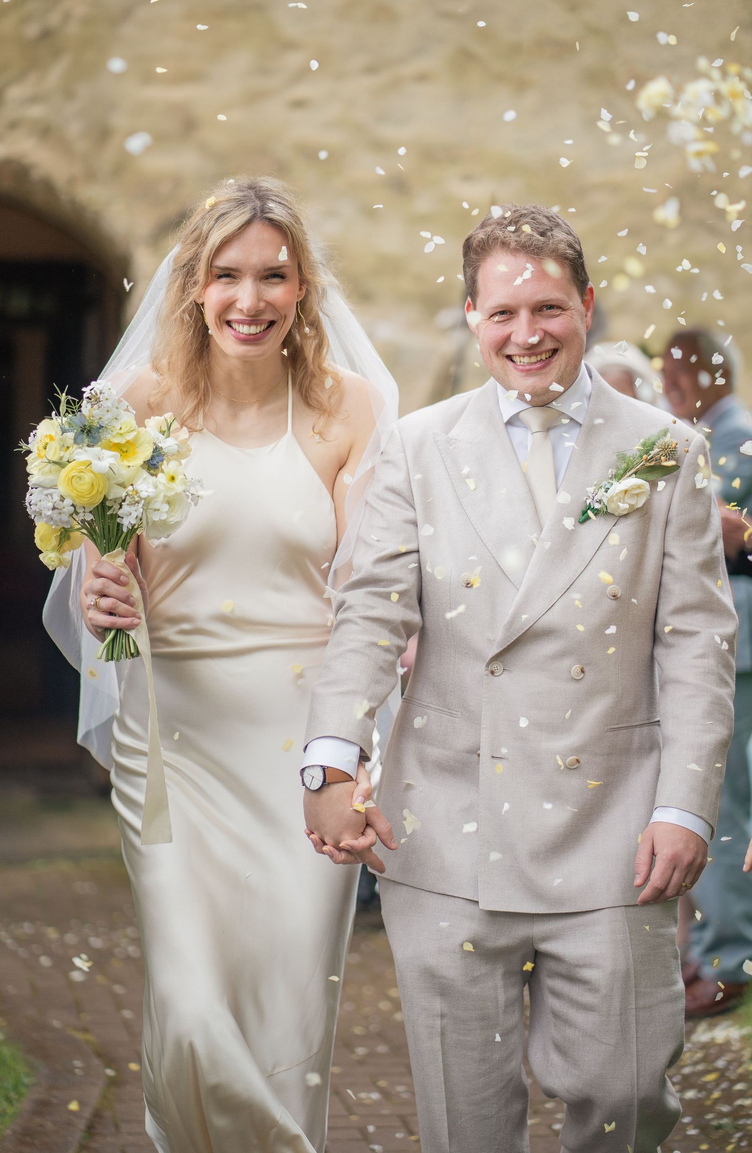 Smiling bride and groom walk hand-in-hand through confetti shower, bride holding yellow bouquet in elegant white gown.