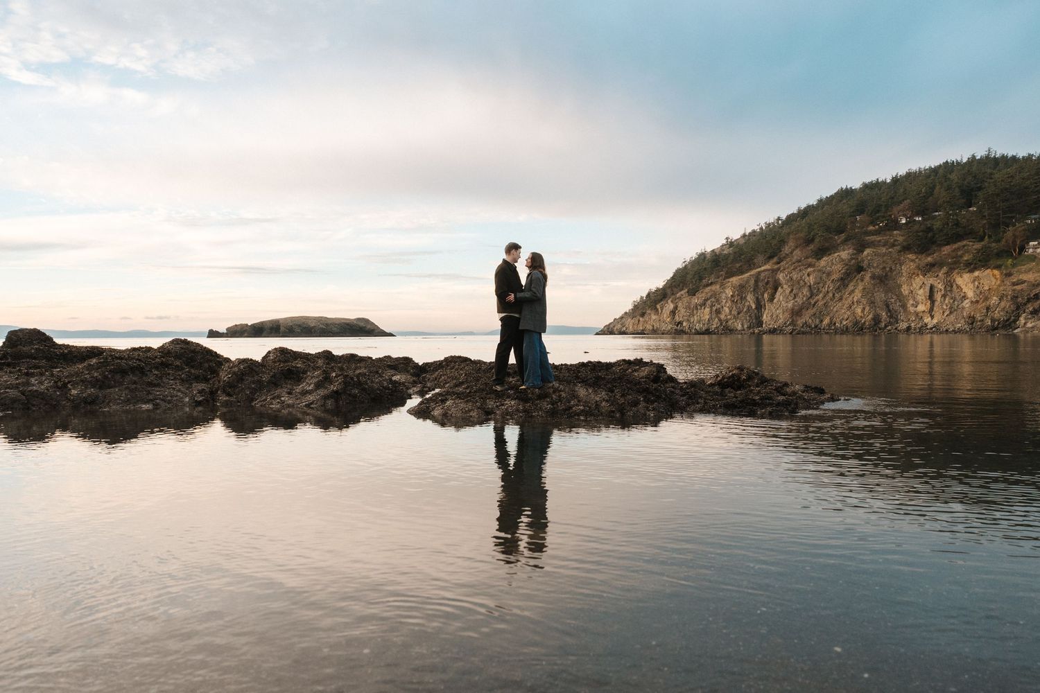 Silhouetted couple stands together at sunset on a rocky beach with mountains and calm water in the background.