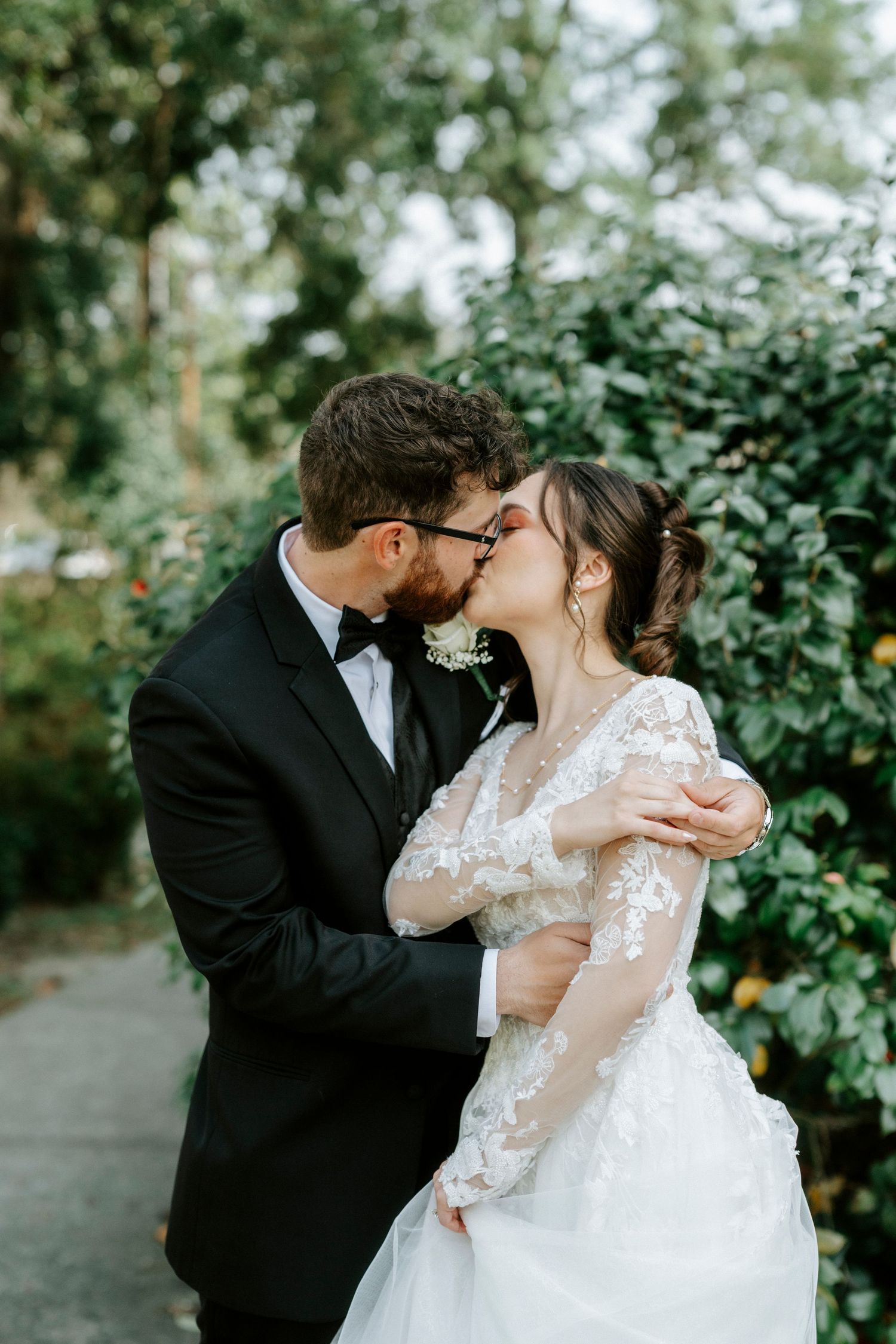 A romantic wedding couple shares intimate kisses in a garden setting with lush greenery in the background.