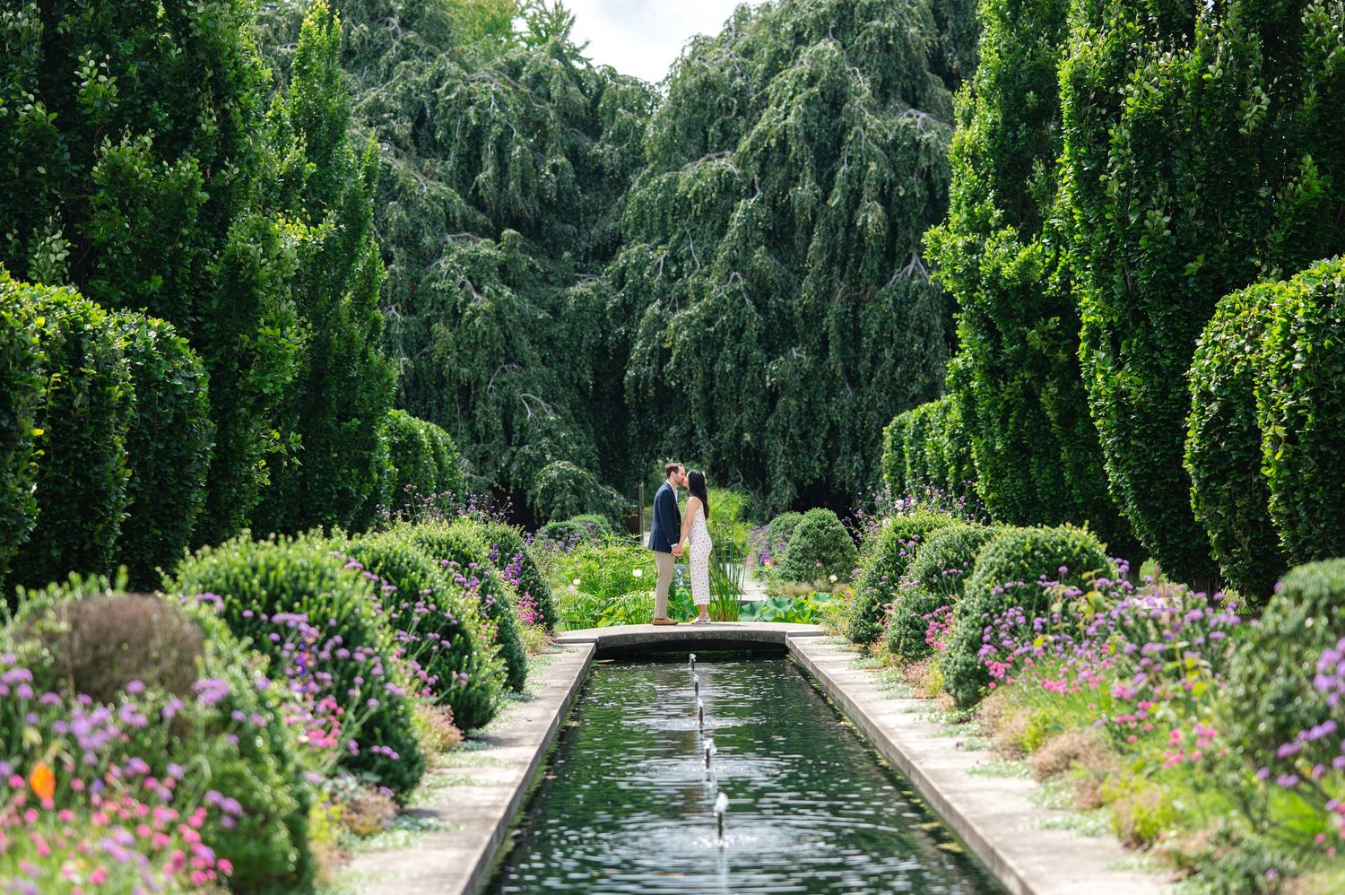 Couple sharing a romantic moment in a lush green garden pathway, woman in floral dress and man in navy suit.