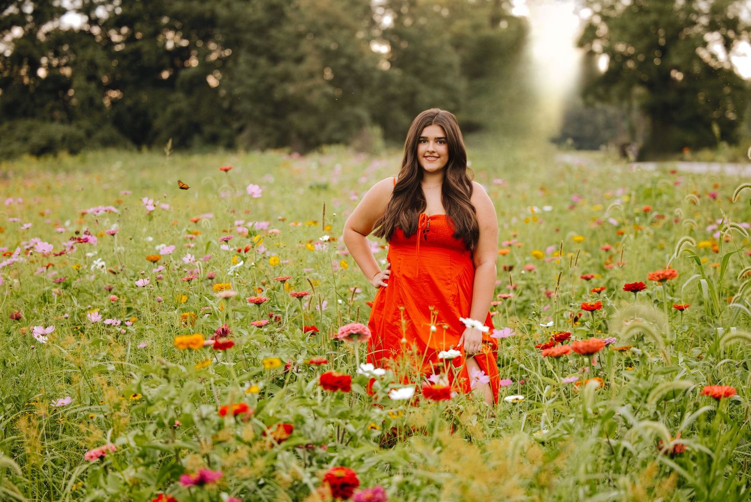 Woman in red dress standing in colorful wildflower field with lush green trees in background.