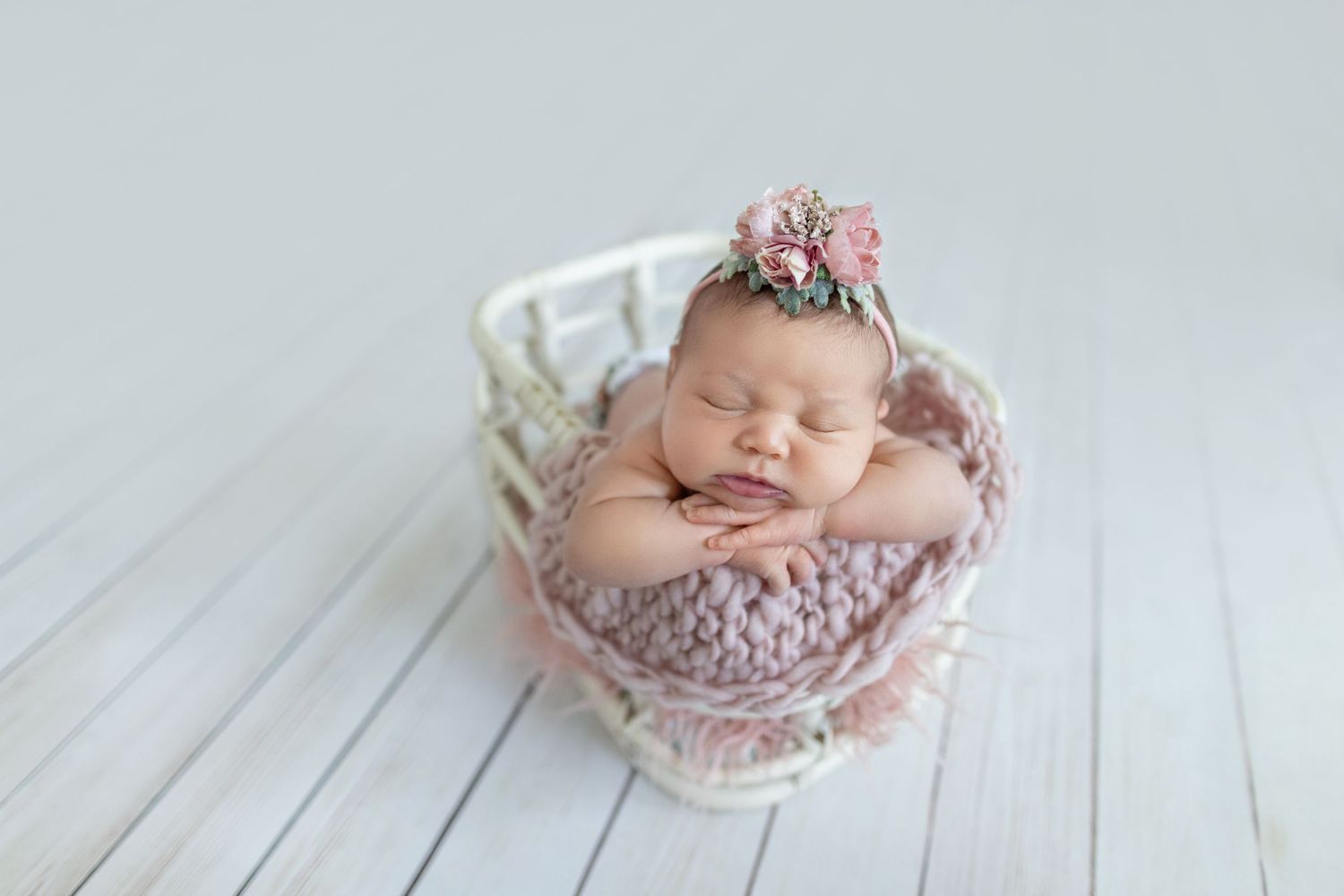 Sleeping newborn baby girl in white basket with pink knit blanket and floral headband on white wood floor.
